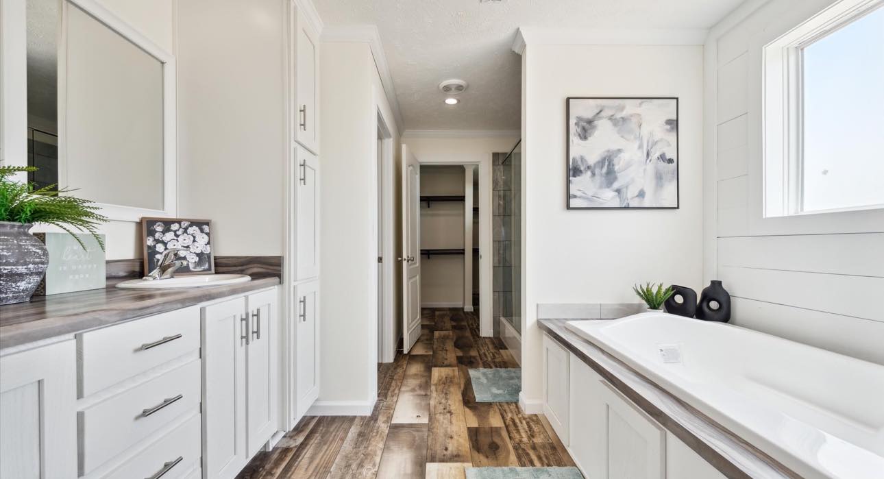 Bright, modern bathroom with wood flooring, white cabinets, and a large tub. Decor includes abstract art, green plants, and a tabletop display.