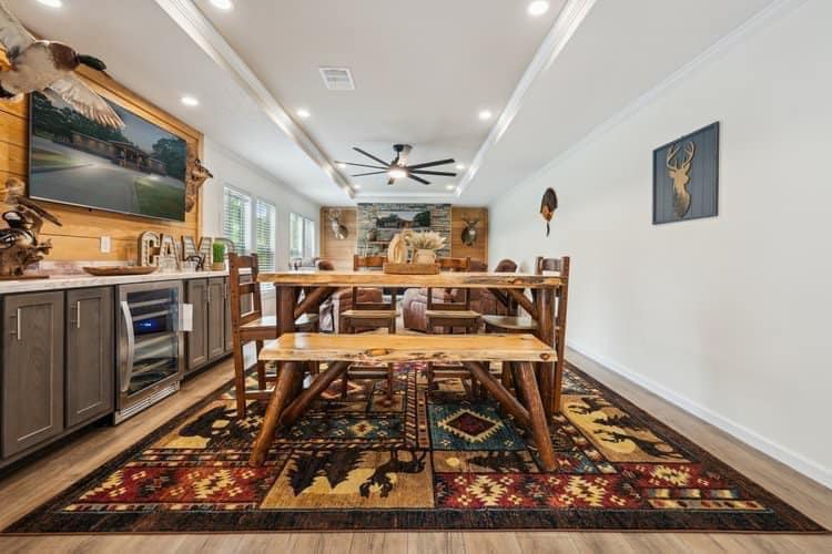 Cozy dining room with rustic wooden table and benches on a patterned rug. Mounted animal decor, landscape art, and sleek ceiling fan enhance a warm, inviting atmosphere.