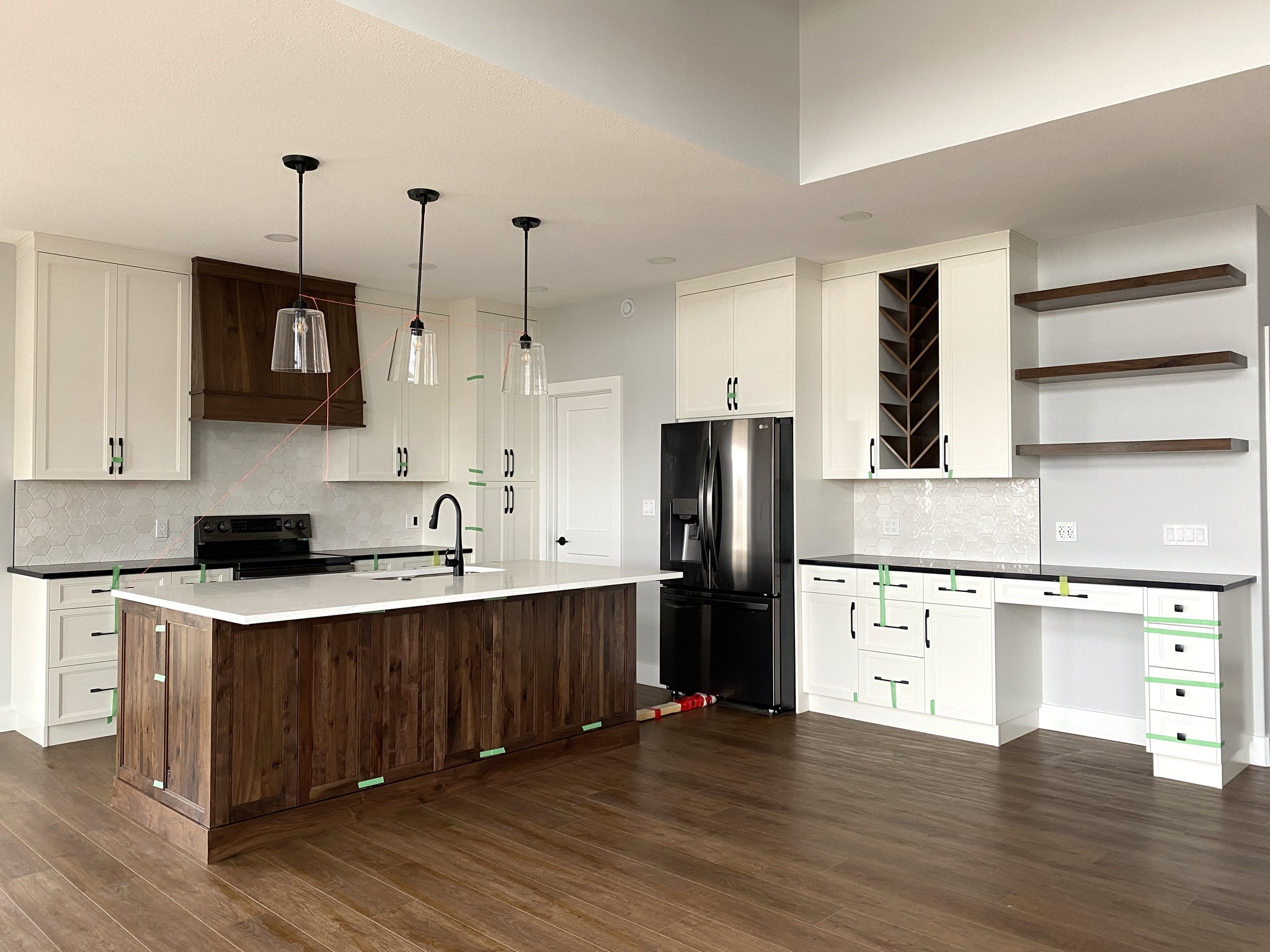 Modern kitchen with dark wood island, white countertops, and cabinets. Black stove and fridge. Three pendant lights hang above. Warm, minimalist design.