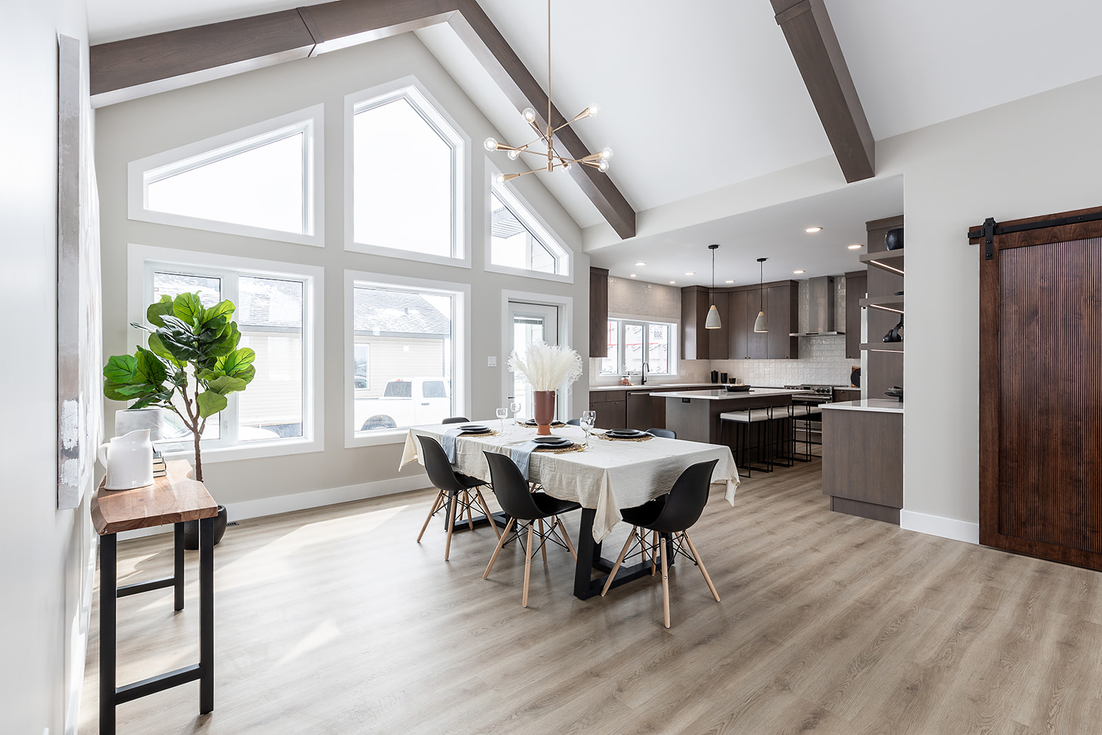 Modern dining room with large angled windows, wooden beams, and a sleek kitchen in the background. A table set for six, and a sliding barn door. Bright and airy.