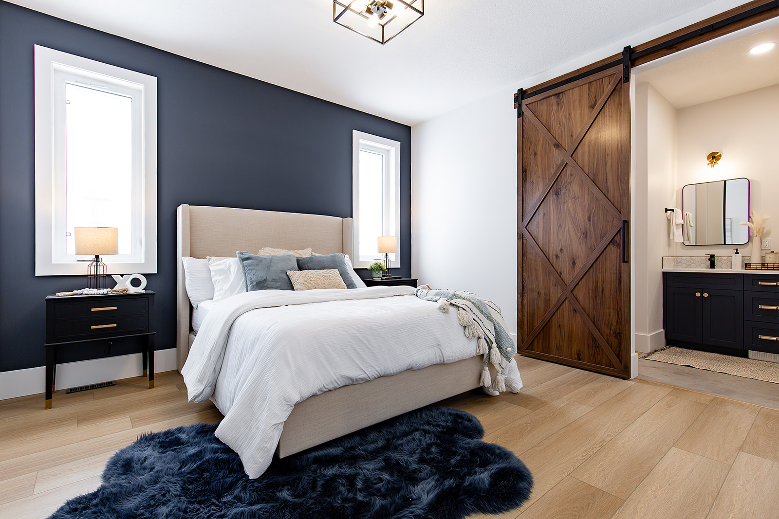 Modern bedroom with a beige bed against a navy accent wall, flanked by two windows. Features wooden sliding barn door, plush rug, and cozy lighting.
