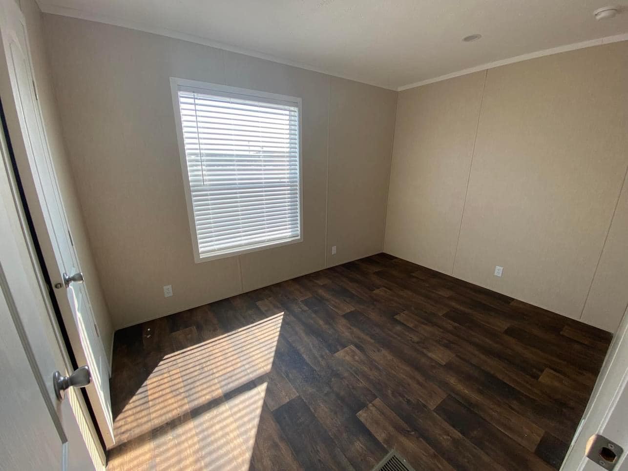 Empty room with beige walls and dark wood flooring. Sunlight streams through a window with closed blinds, casting shadows on the floor. Minimalist and calm.