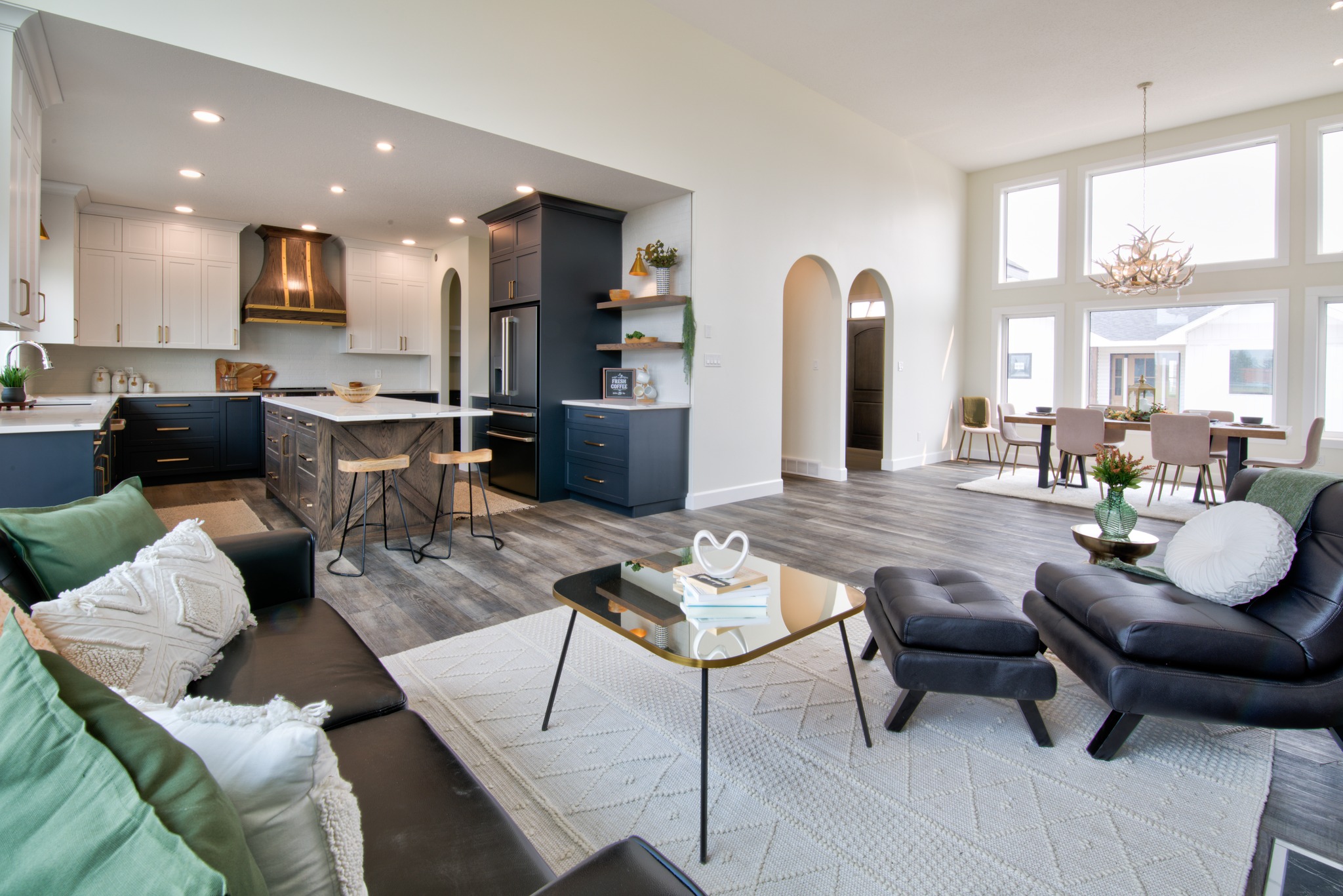 Spacious open-plan living area with modern kitchen, dining, and lounge. Features dark cabinets, wooden stools, light-filled windows, and cozy seating.