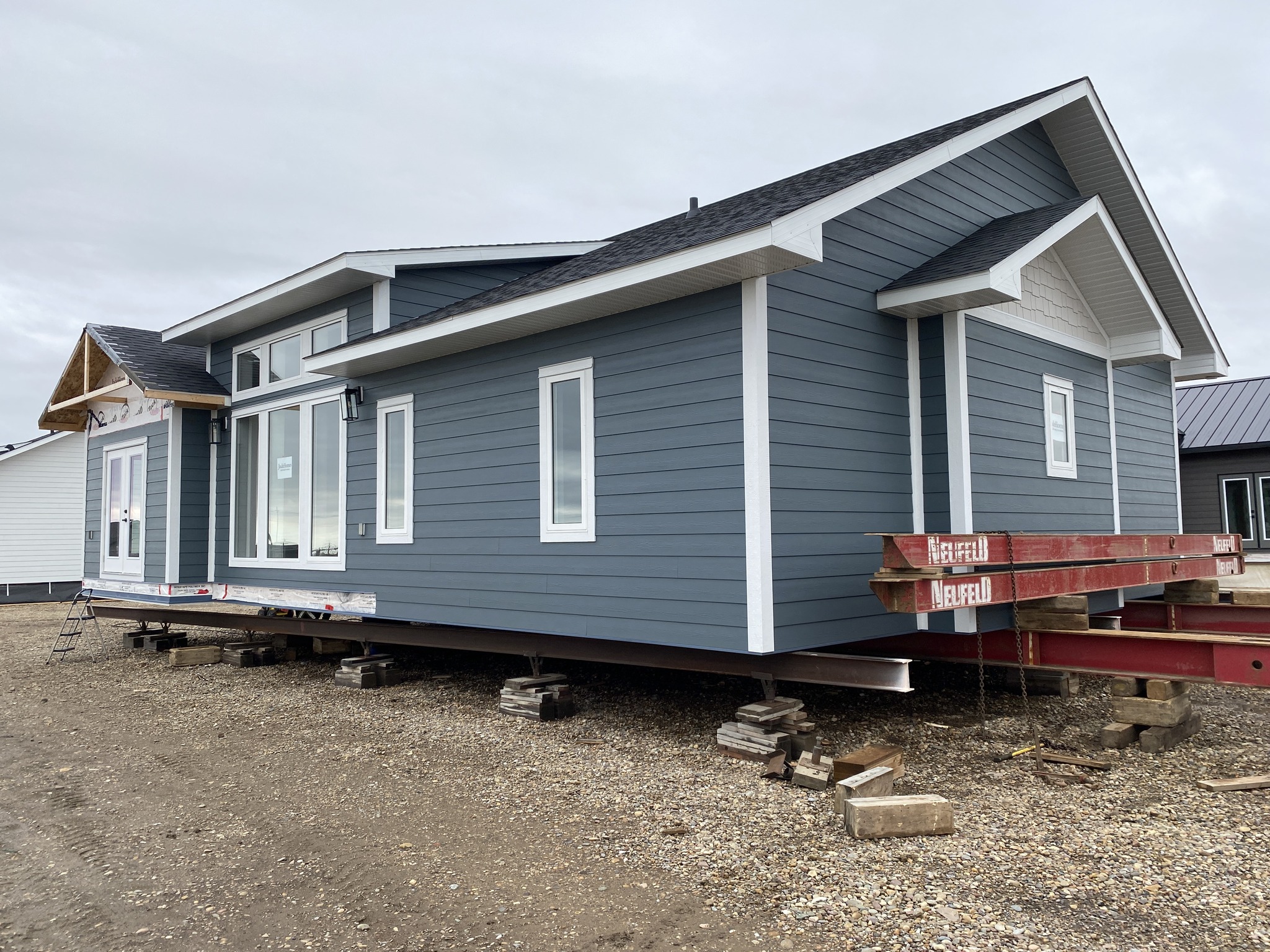 A modern, blue-gray modular home elevated on blocks, featuring white trim and large windows, sits on a gravel lot under an overcast sky.