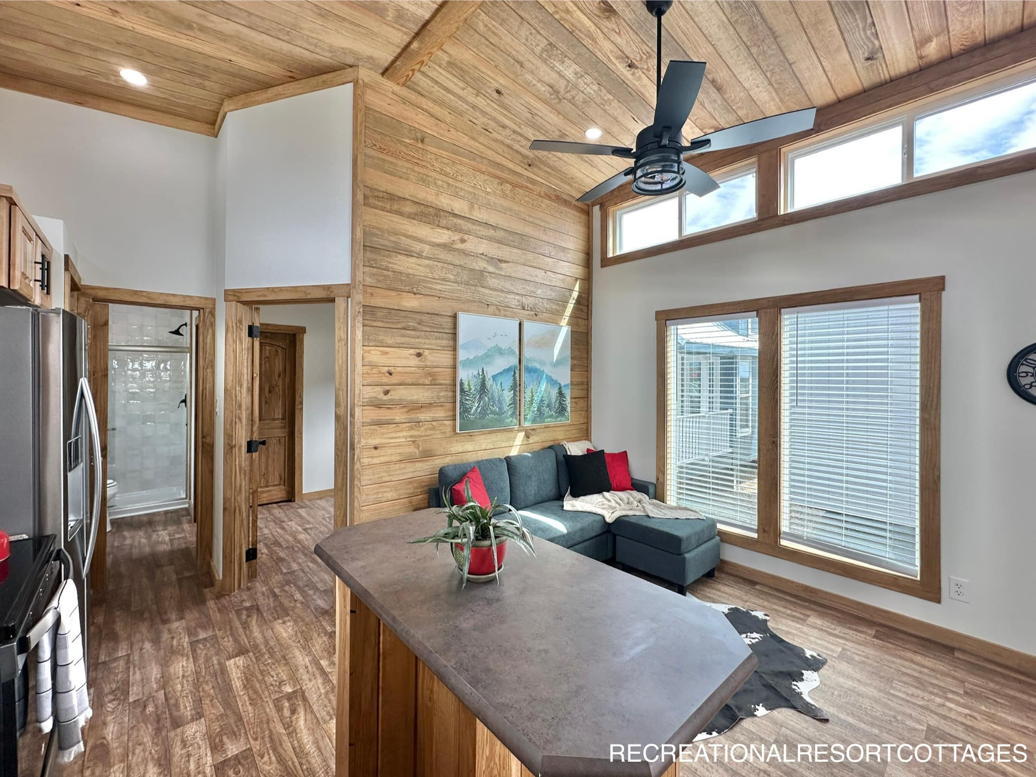 Stylish tiny home interior with wood paneling, vaulted ceiling, and large windows. Features a gray sectional sofa, modern kitchen island, and ceiling fan.