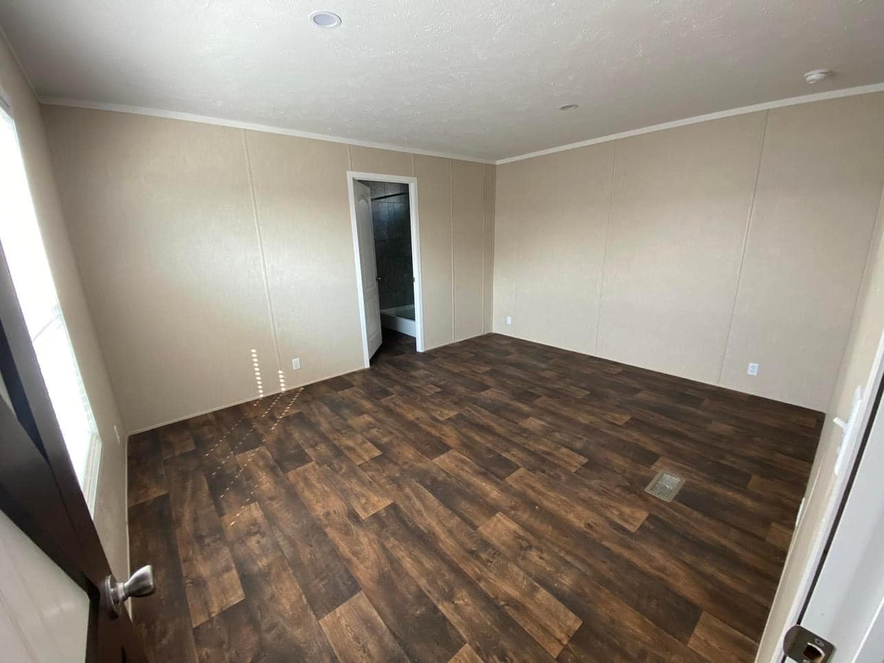 Empty room with beige walls and dark wood flooring. A door on the far wall opens to a bathroom. Sunlight streams in from a window, creating a calm atmosphere.