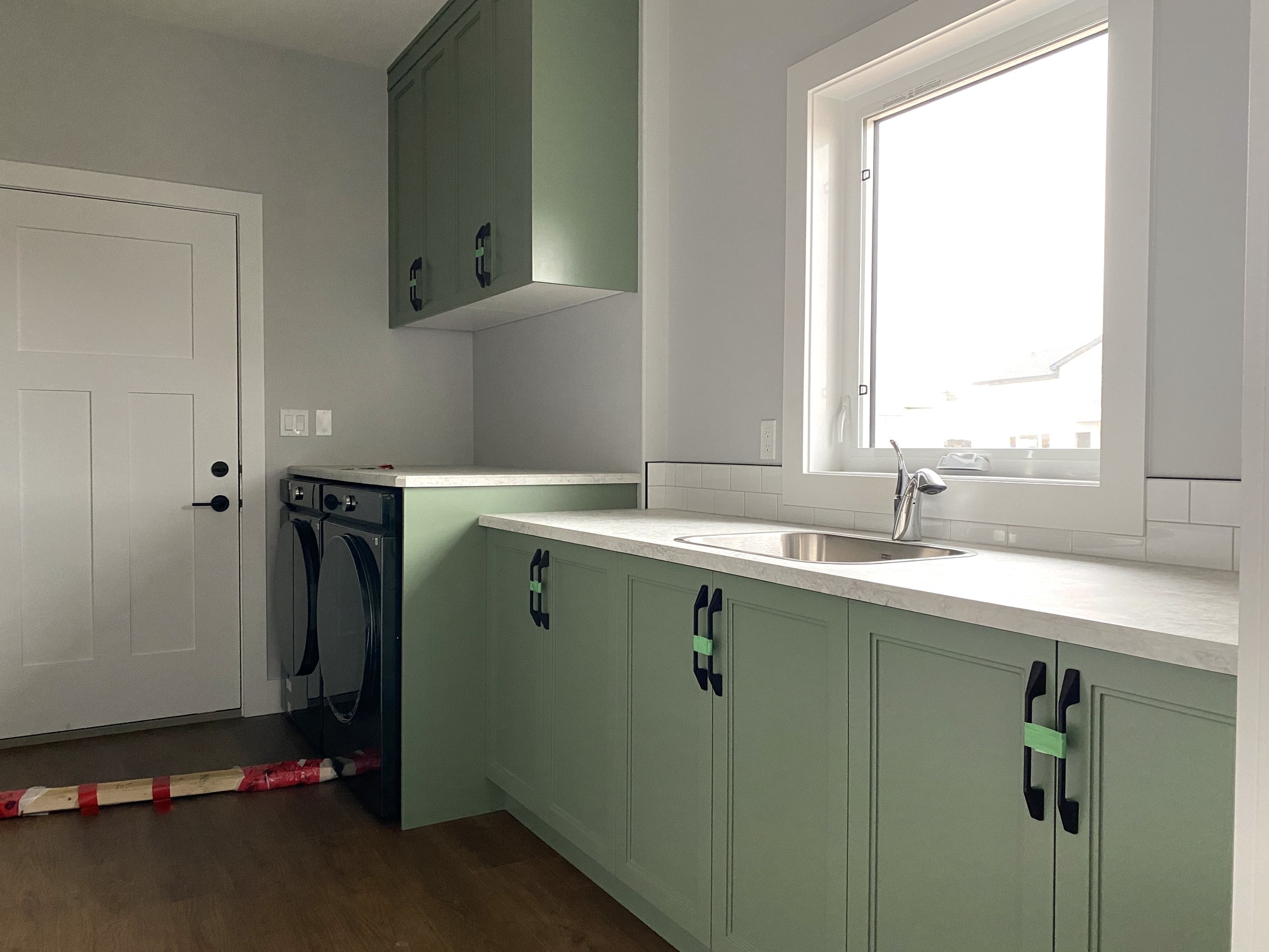 A laundry room with light green cabinets, black handles, and a white countertop. There is a silver sink below a window on the right. A washer and dryer are set on the left near a door. The room has a modern, clean feel.