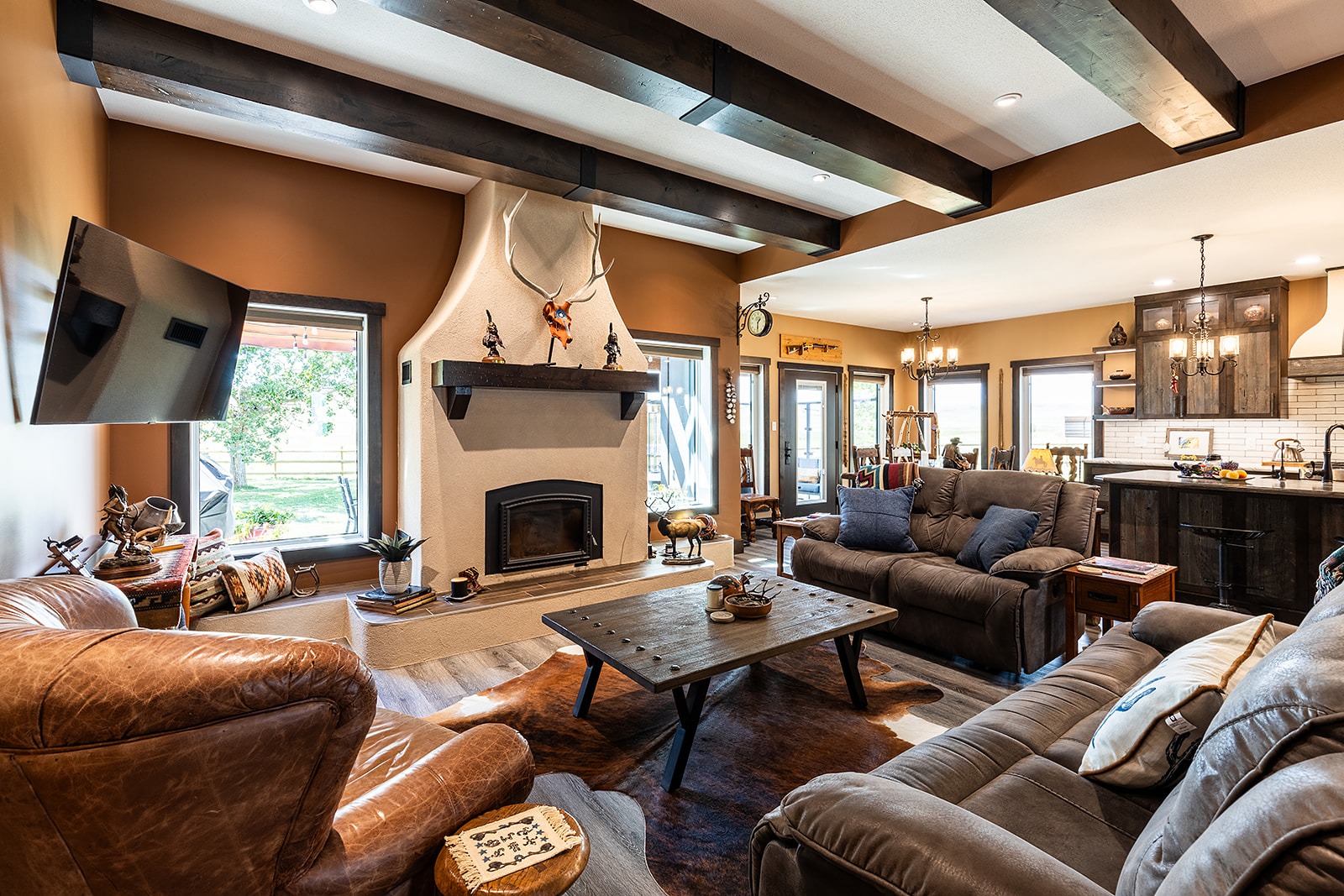 Rustic living room with wooden beams and warm tones. Features a cozy sofa, leather armchair, fireplace with deer antlers, and a wooden coffee table.