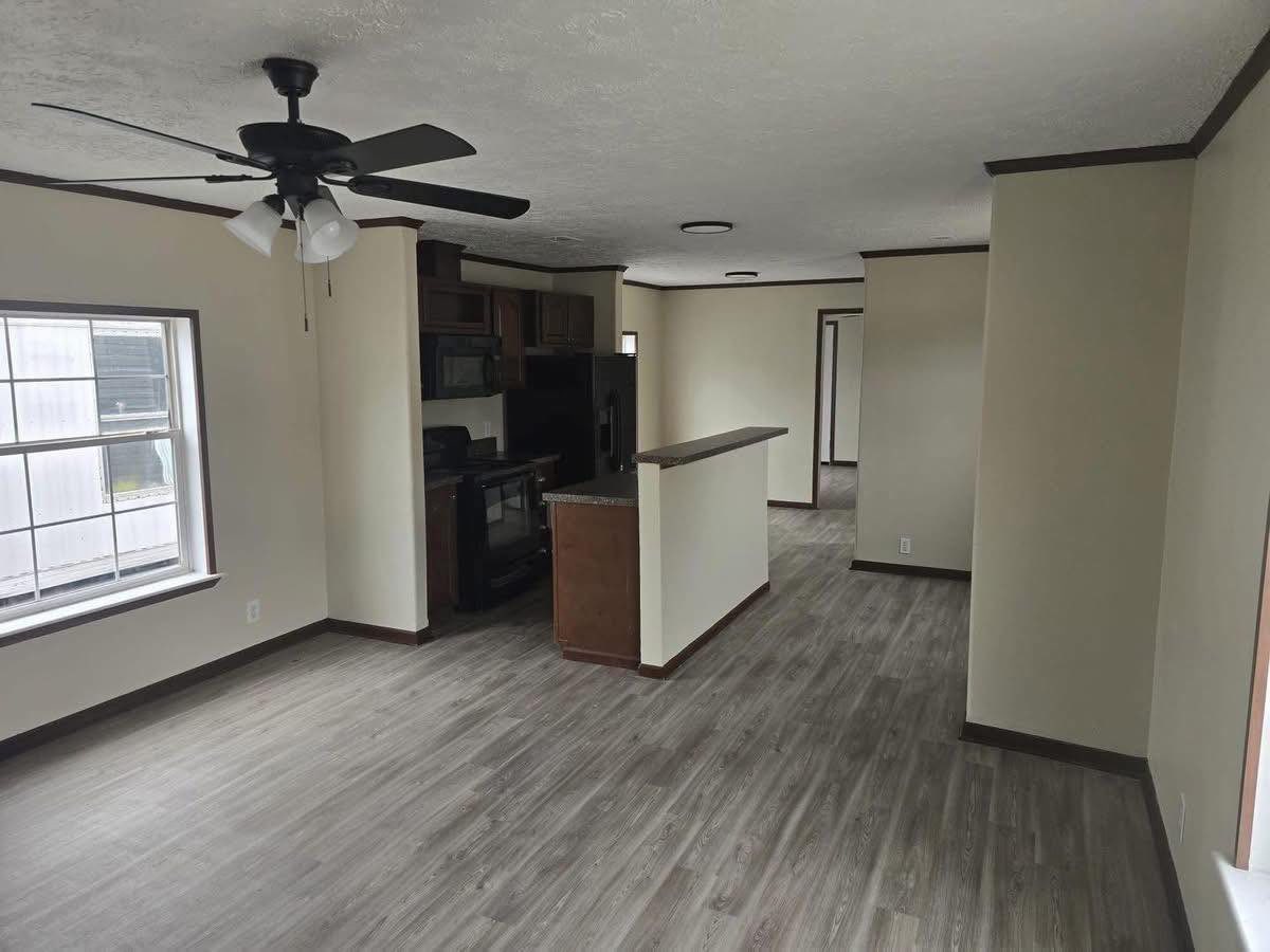 Spacious room with wood flooring, beige walls, and a ceiling fan. Features include a kitchen with dark cabinets, a window, and a partition wall.