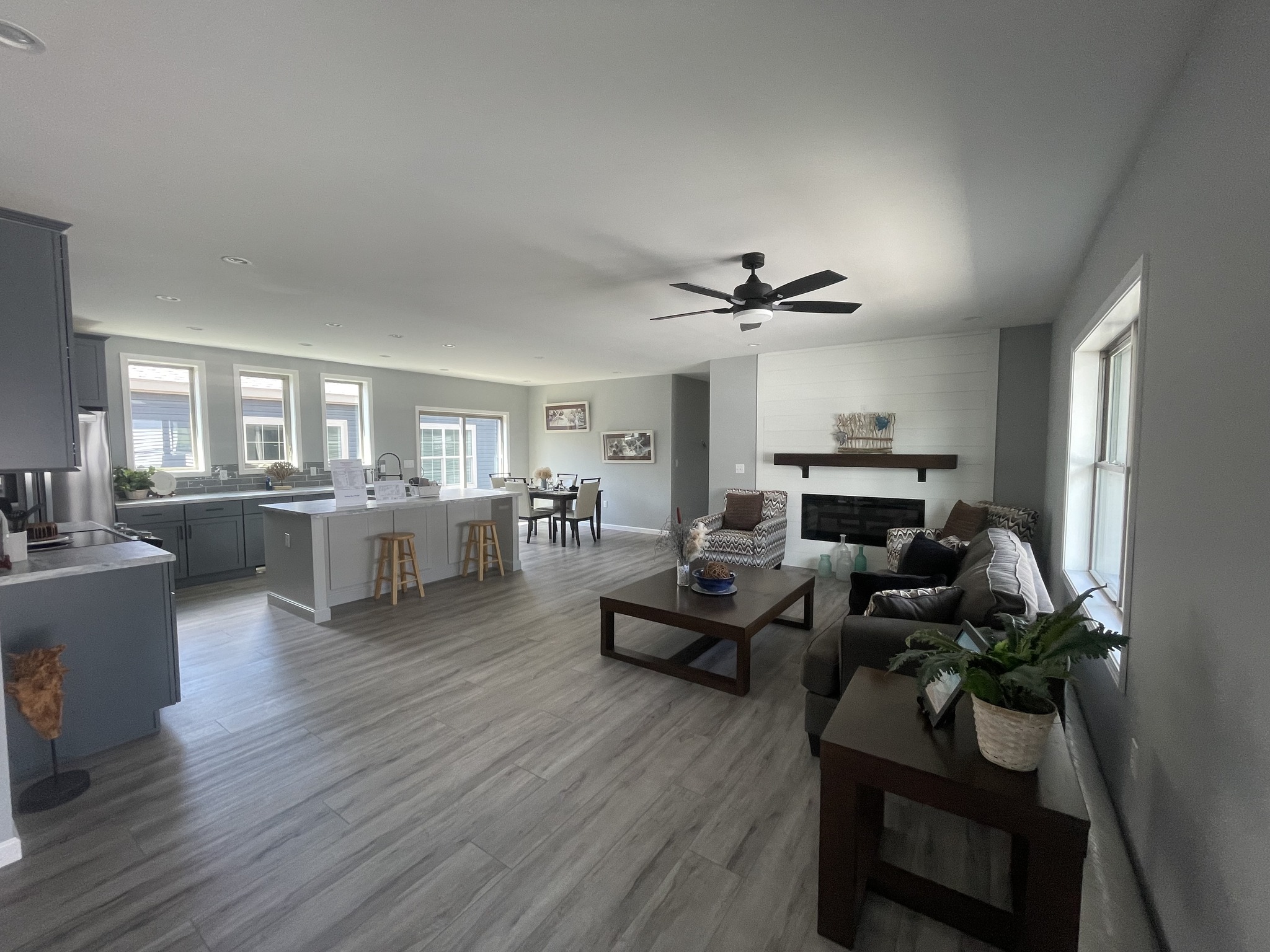 Spacious open-plan living area with gray flooring, featuring a modern kitchen and cozy seating by a fireplace. Bright windows and minimalist decor.