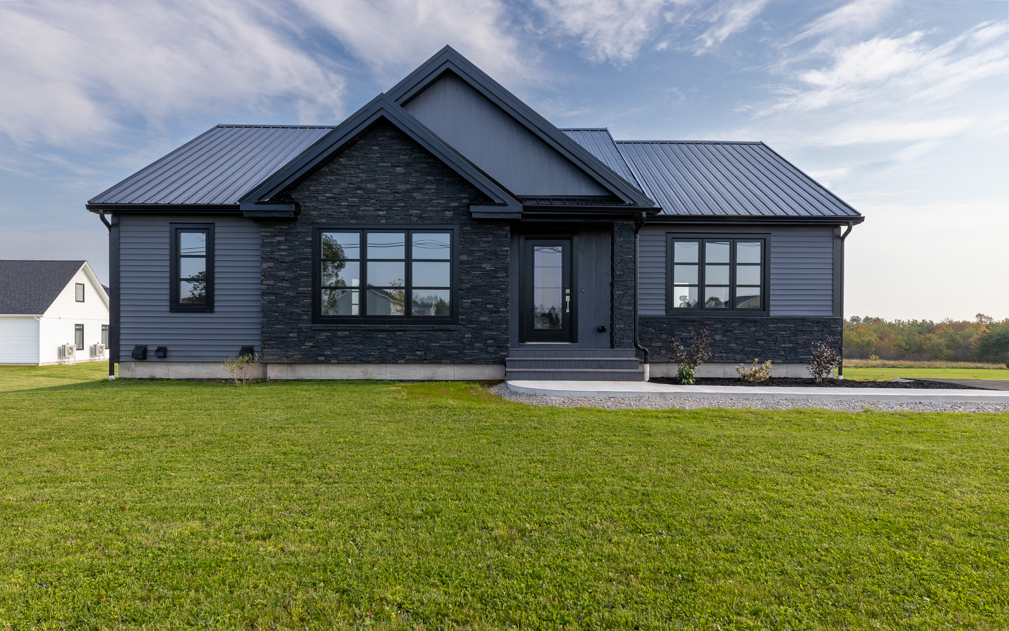 A modern, single-story house with dark gray siding and large windows, set on a neatly manicured lawn. The sky is partly cloudy, creating a calm atmosphere.