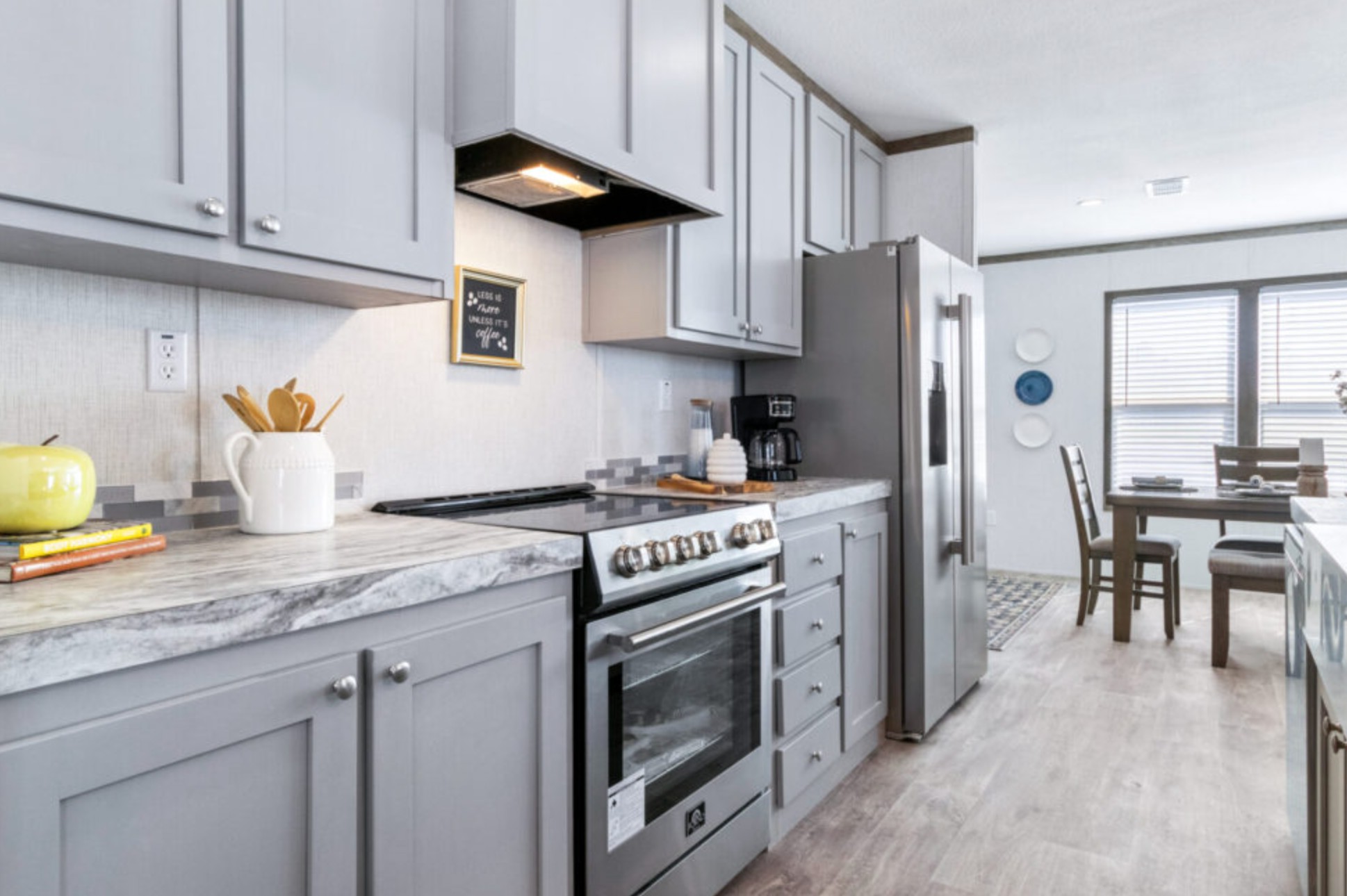 Modern kitchen with gray cabinets, marble countertops, and stainless steel appliances. A dining table is visible in the background. Bright and spacious.
