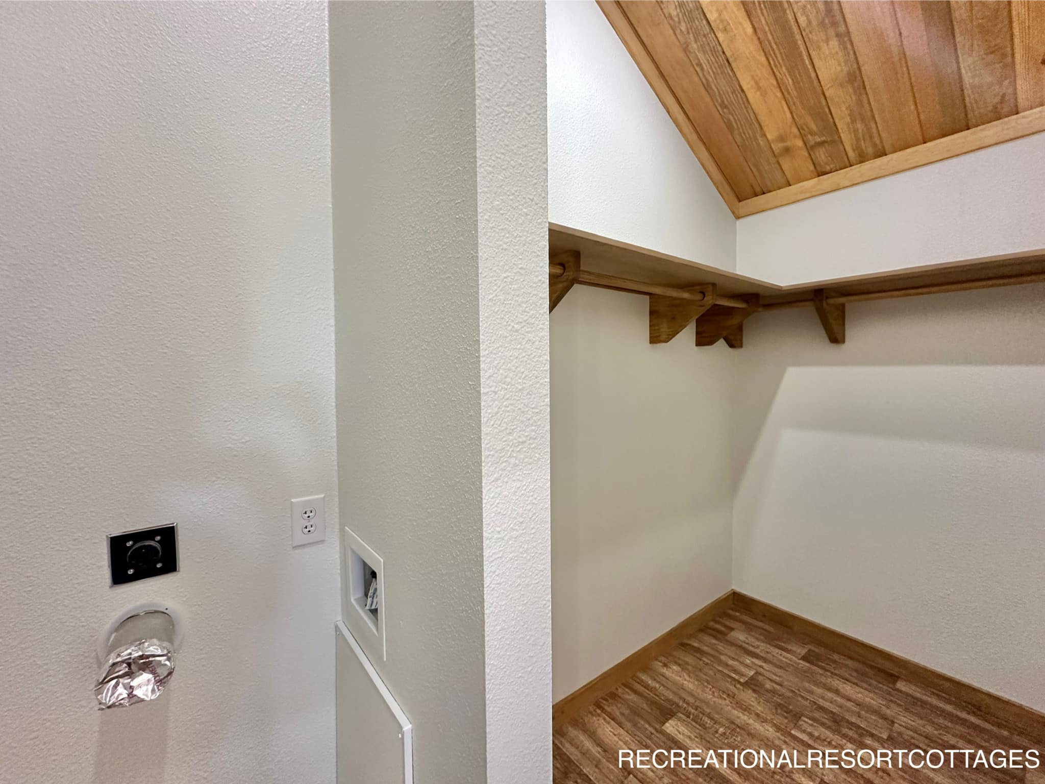 Wood-paneled ceiling and laminate floor in a small, empty room. Wall outlet and vent on the left; wooden shelf wraps around two walls, creating a cozy feel.