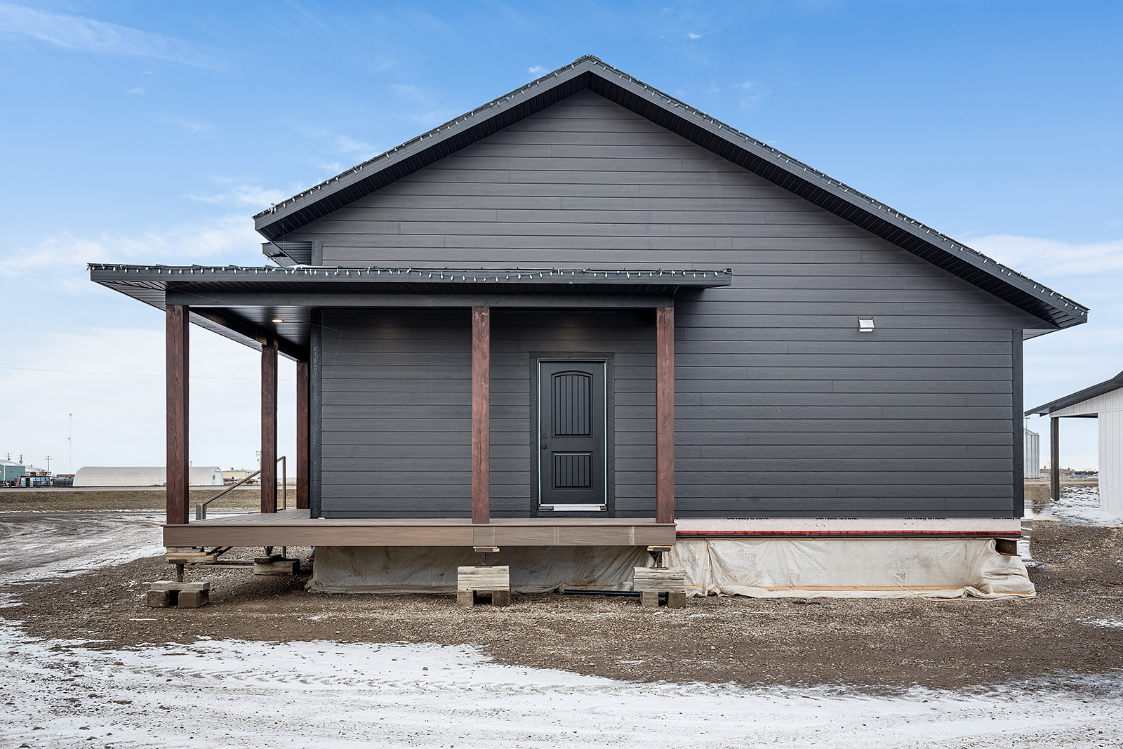 A modern, dark gray house with a wooden porch and triangular roof set against a clear blue sky. Snow and dirt surround the elevated structure.