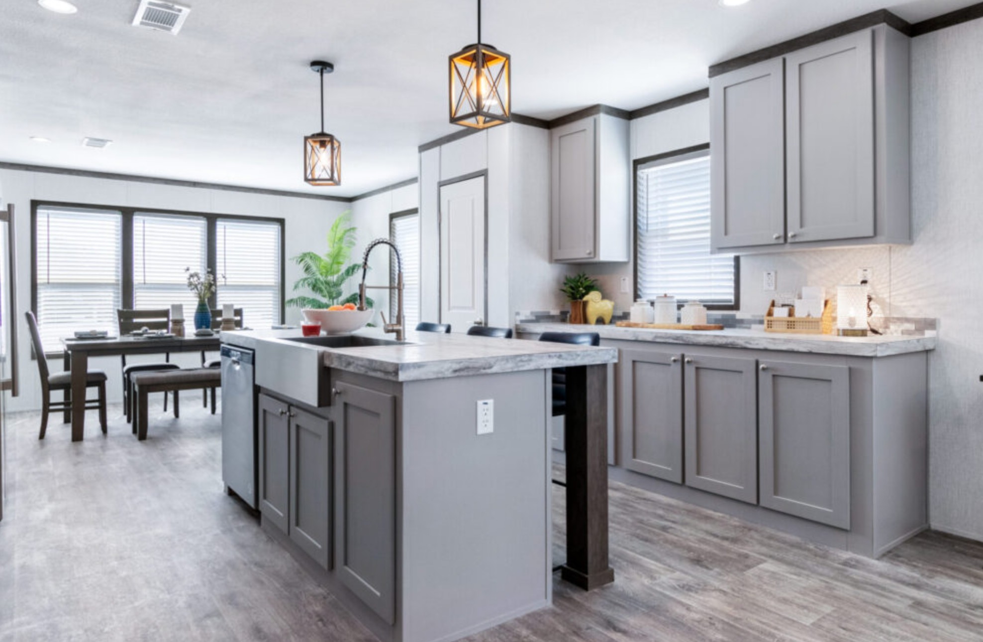 Modern kitchen with gray cabinets, marble countertops, and island. Hanging pendant lights add warmth. Dining area with large windows in background.