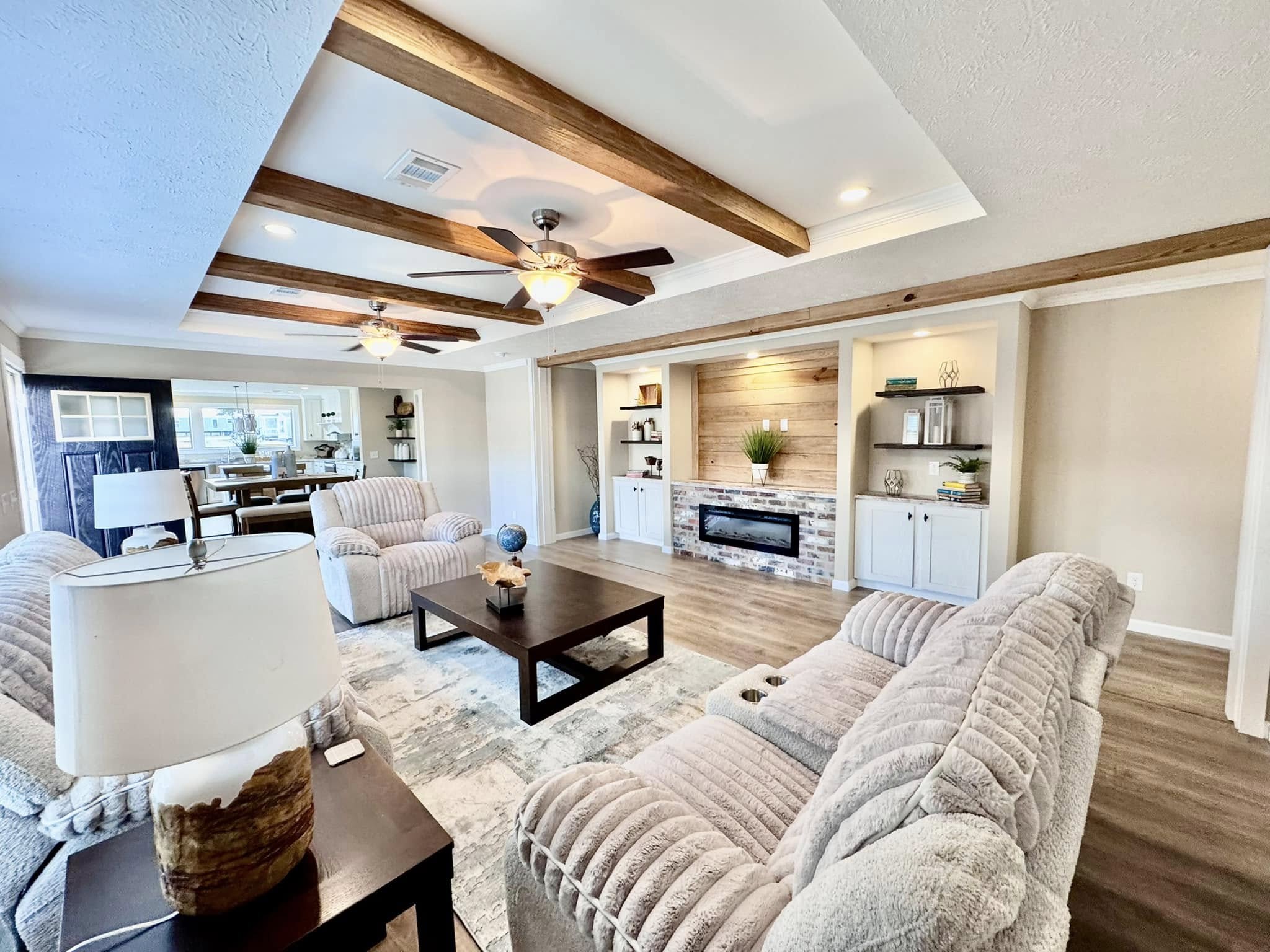 Spacious living room with beige walls, ceiling beams, and ceiling fans. Features plush sofas, a dark wood coffee table, and a modern fireplace.