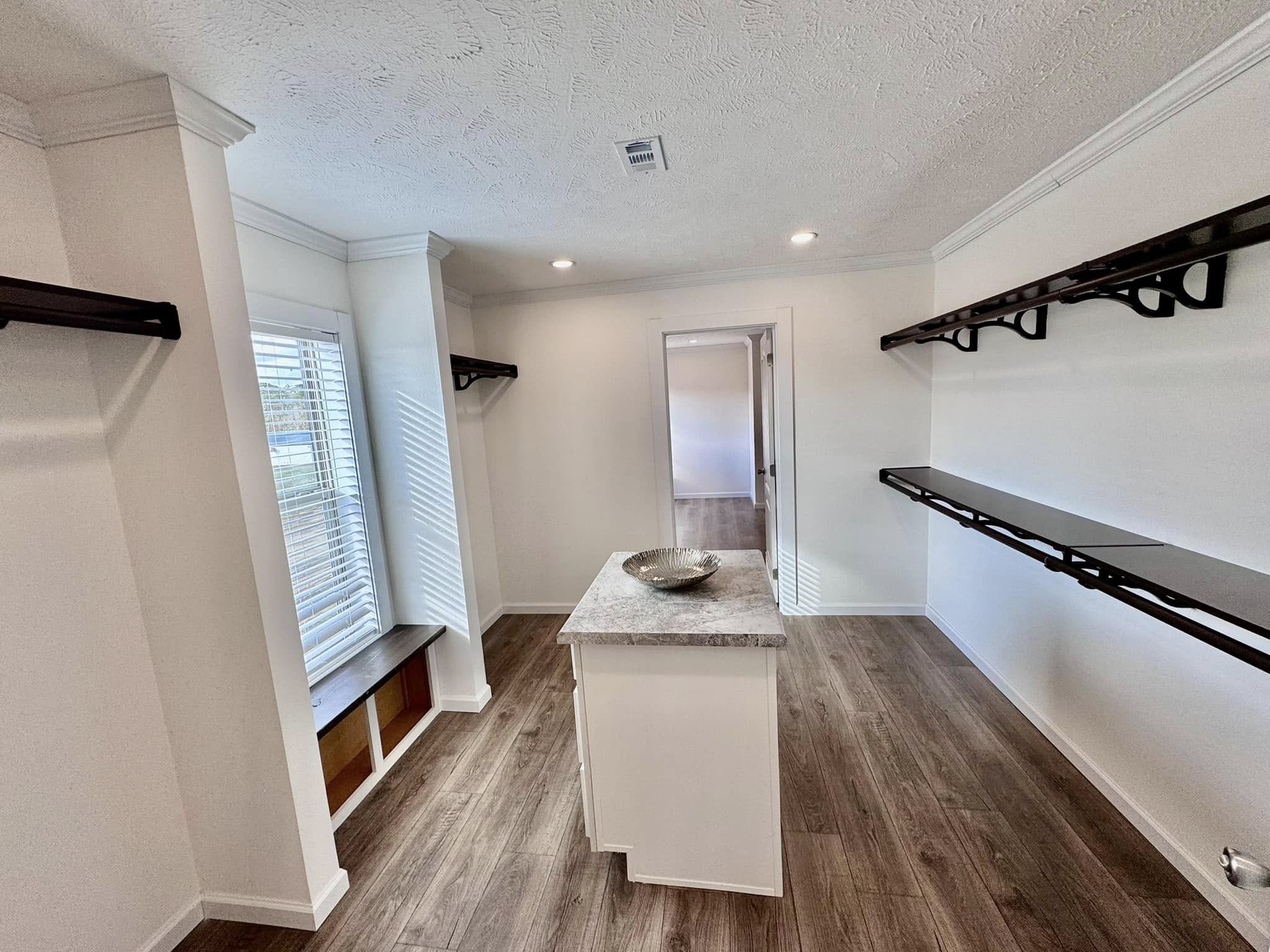 Bright walk-in closet featuring wooden flooring, long dark shelves on the right, a small island with a decorative bowl, and a window bench on the left.