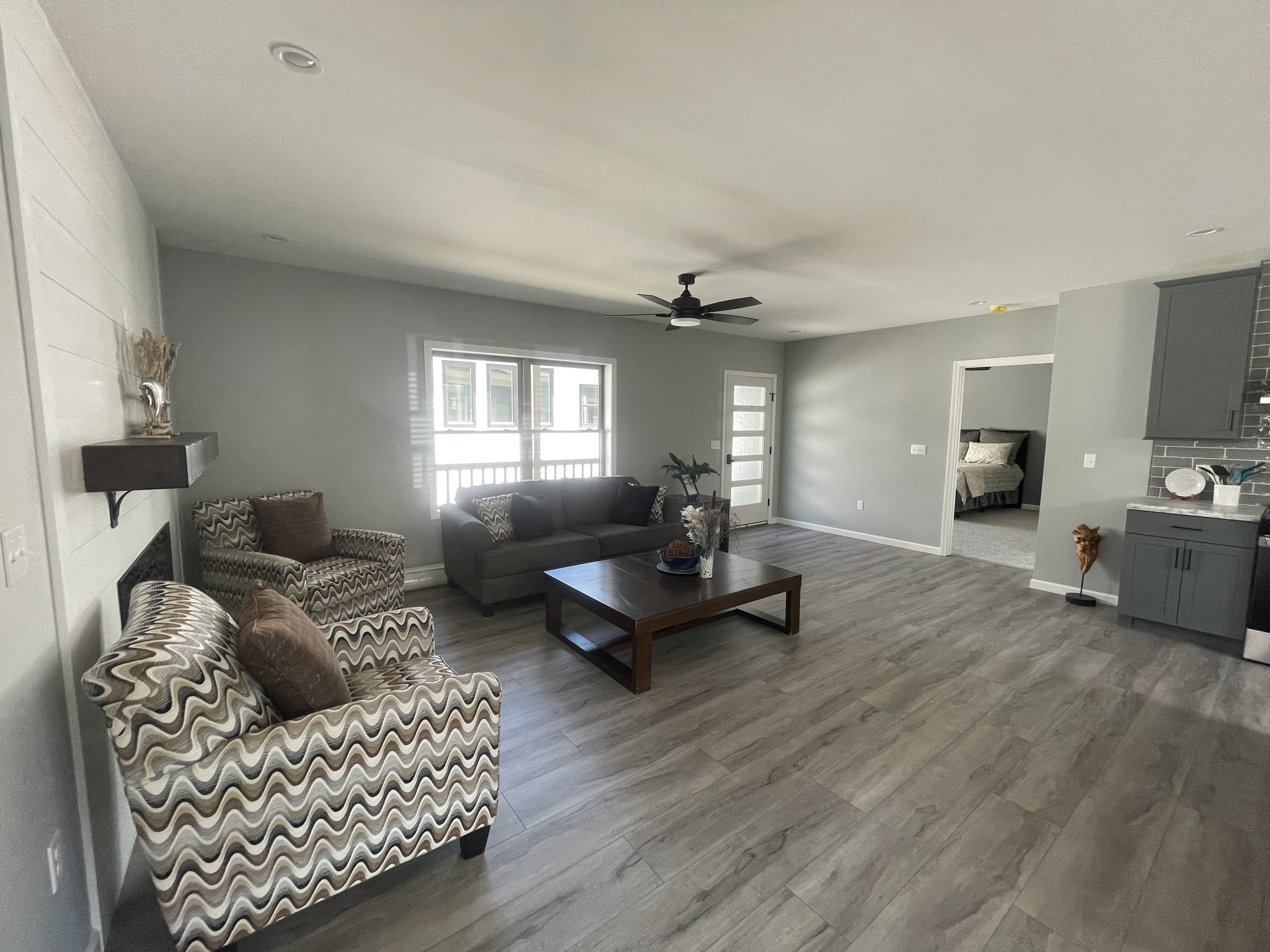 Spacious living room with gray walls and wood flooring. Features a brown sofa, patterned armchairs, a large coffee table, and a ceiling fan. Natural light streams through a window, creating a cozy ambiance.