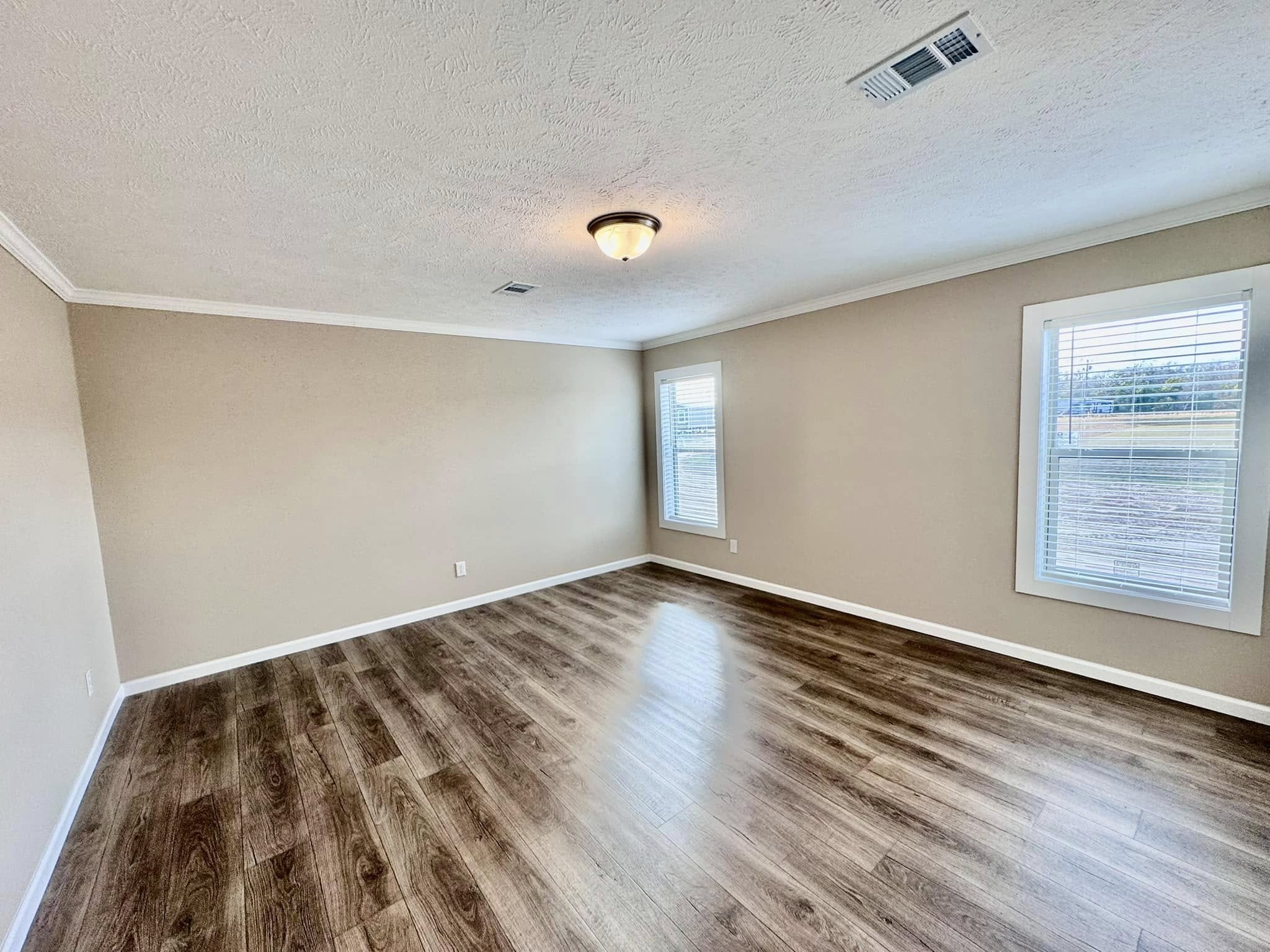 A spacious, empty room with light brown walls and dark wood flooring. Two windows allow natural light in, creating a calm, open atmosphere.