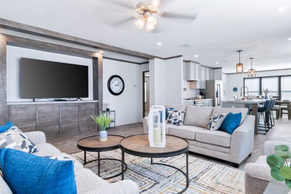 Modern living room with gray sofas, blue cushions, and round coffee tables on a patterned rug. Wall-mounted TV, ceiling fan, and open kitchen in the background. Cozy and inviting atmosphere.