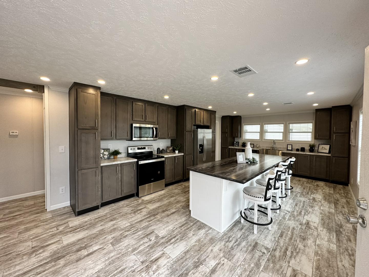 Spacious kitchen with rustic wood flooring, rich brown cabinets, stainless steel appliances, large island with stools, and natural light from three windows.