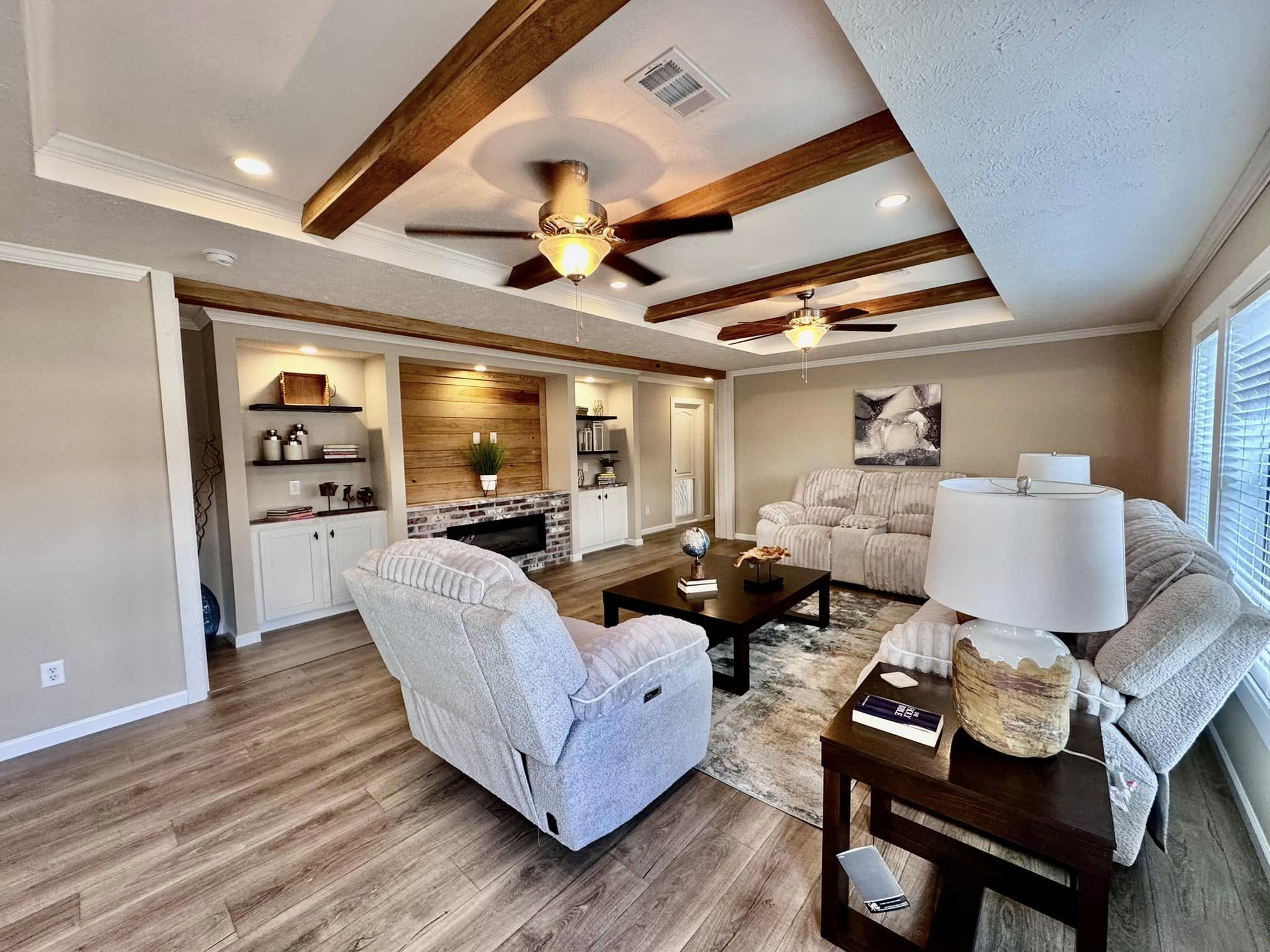 Spacious living room with beige walls, wood beams, plush sofas, and a central coffee table. Cozy atmosphere with ceiling fans and large windows.