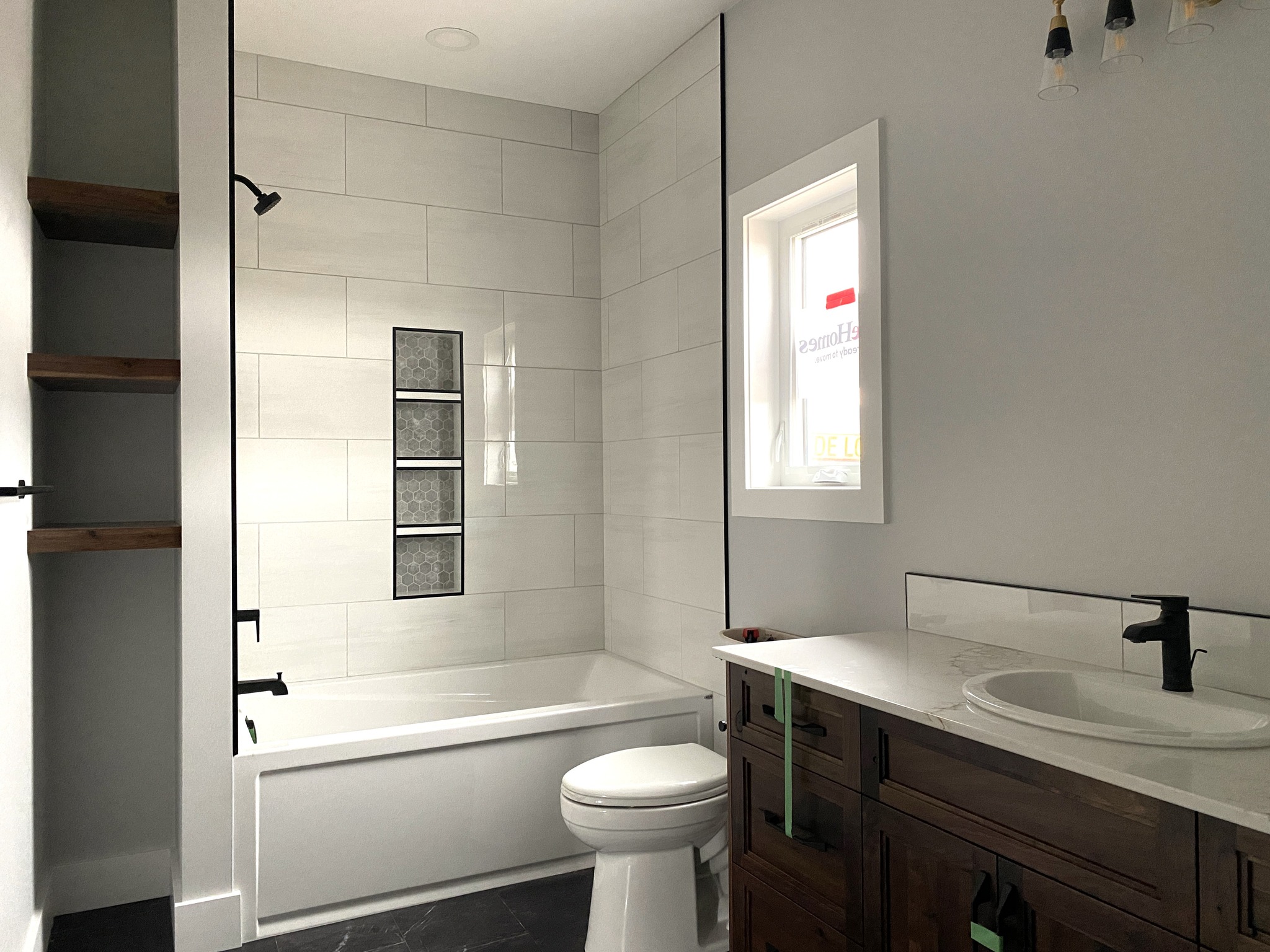 Modern bathroom with light gray tiles and a built-in bathtub. Wooden vanity with white countertop, black fixtures, open shelves, and a small window.