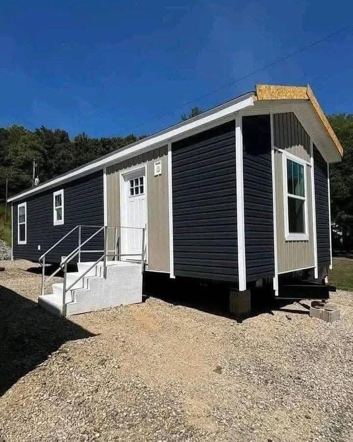 Small, modern mobile home with dark siding and white trim, set on a gravel lot. A short staircase leads to the front door, with trees in the background.