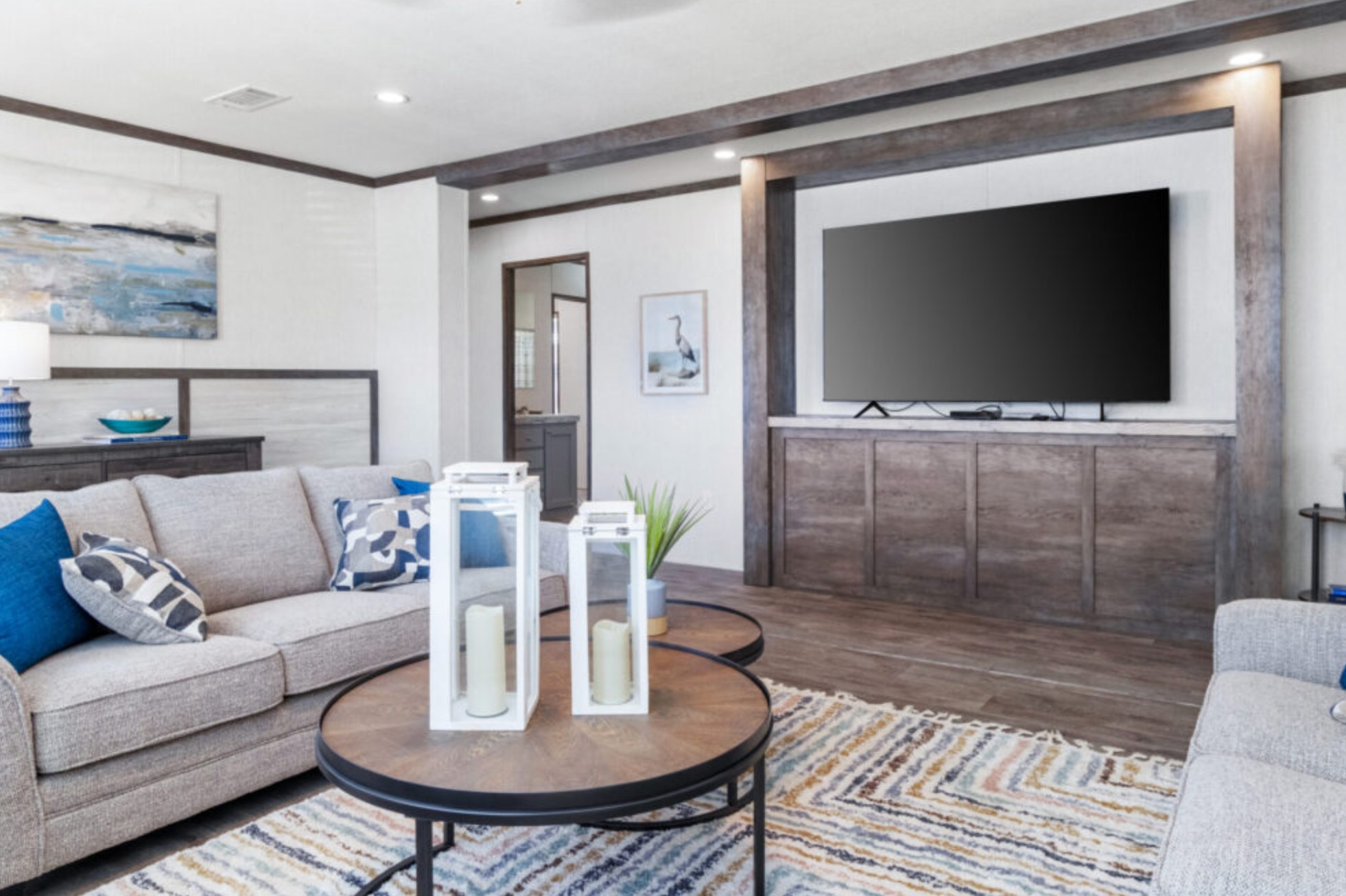 Modern living room with neutral tones. Features gray sofas, blue and patterned cushions, a large TV, and decorative candles on a round coffee table. Cozy and inviting ambiance.