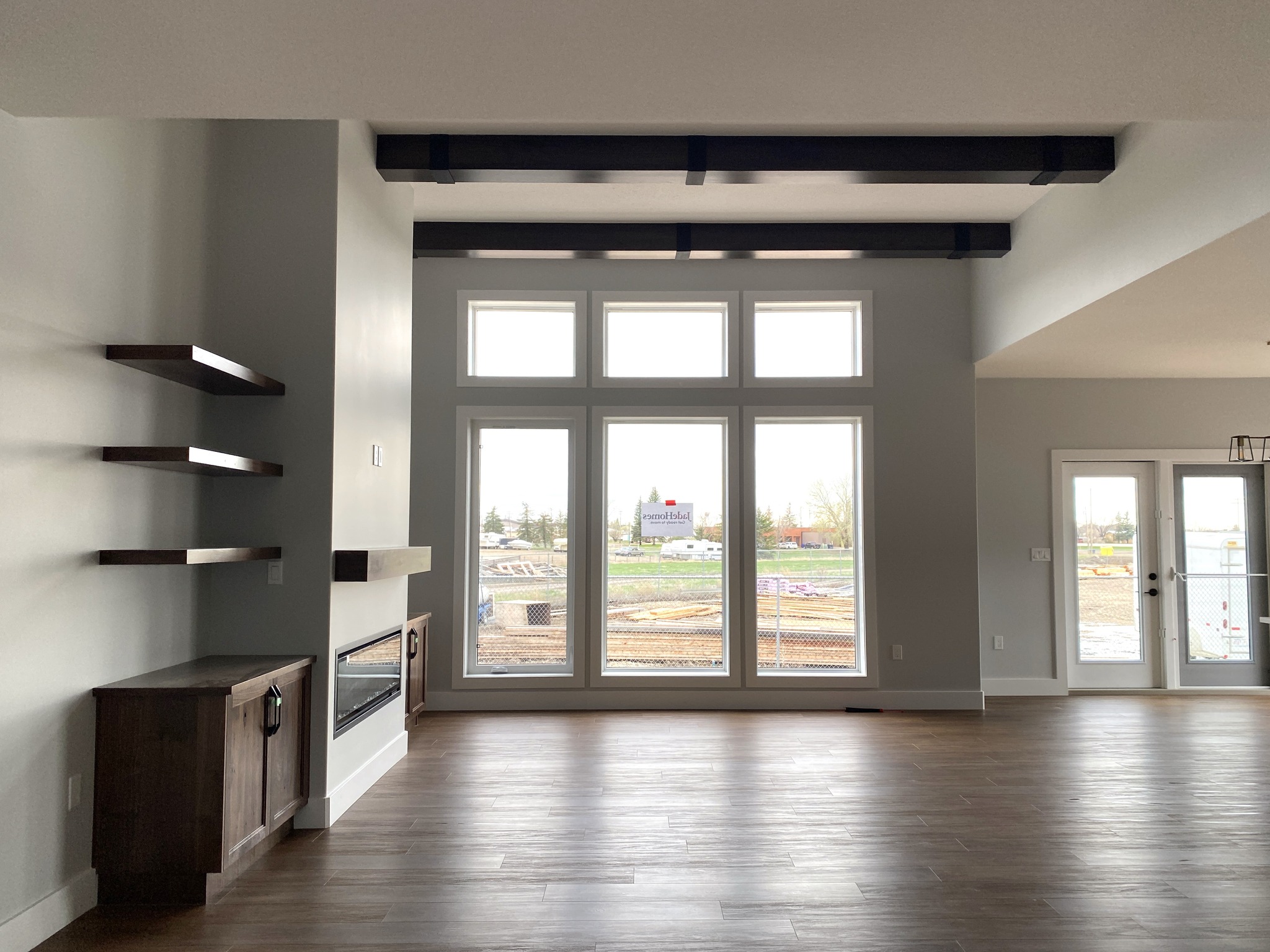 Spacious modern living room with large floor-to-ceiling windows, wood floors, dark ceiling beams, floating shelves, and a fireplace beside a wooden cabinet.