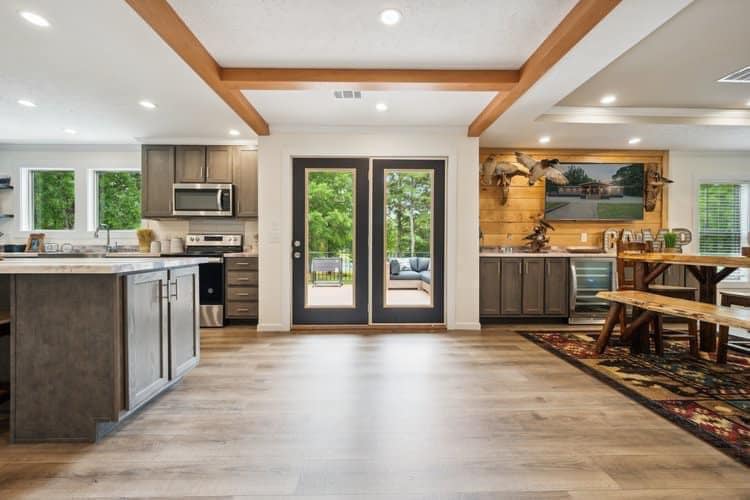 Modern kitchen and dining space with wooden floors, exposed beams, and large glass doors overlooking greenery. Cozy, warm, and inviting ambiance.