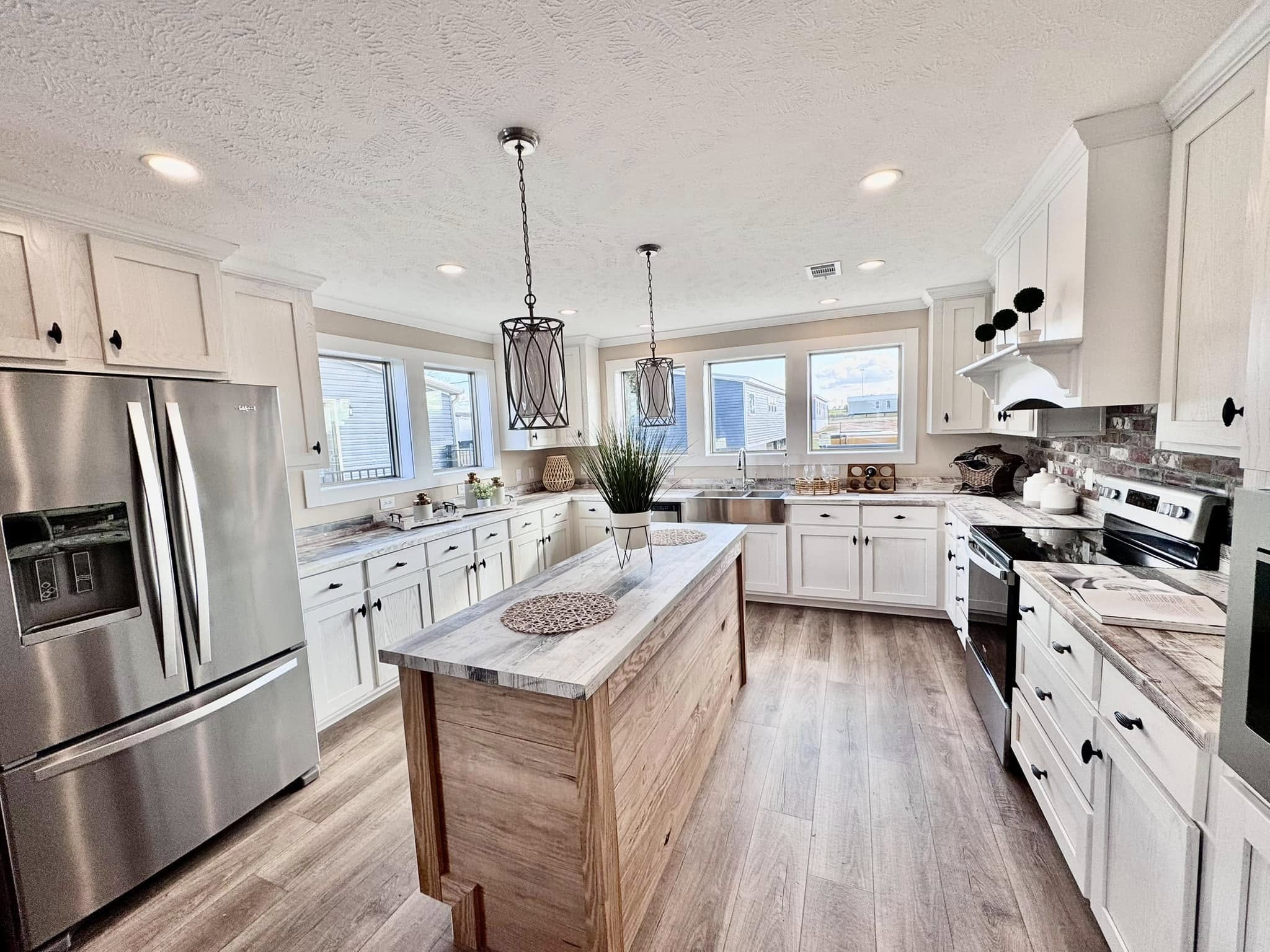 Spacious kitchen with white cabinets, stainless steel appliances, and a wooden island. Pendant lights hang from the ceiling, with large windows providing natural light.