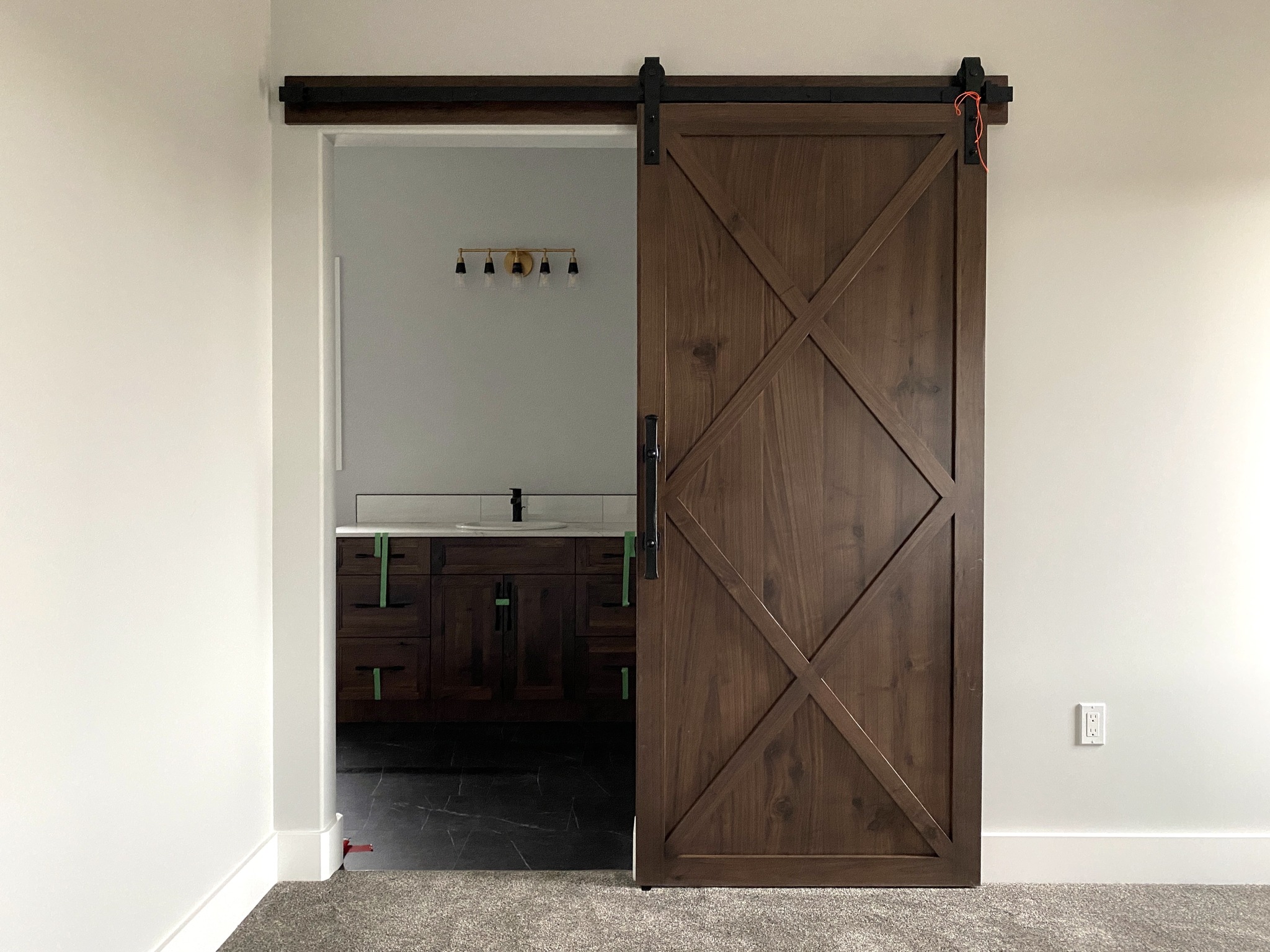 A wooden sliding barn door partially open, revealing a bathroom with a modern sink and dark cabinetry. Soft lighting and a minimalist style.
