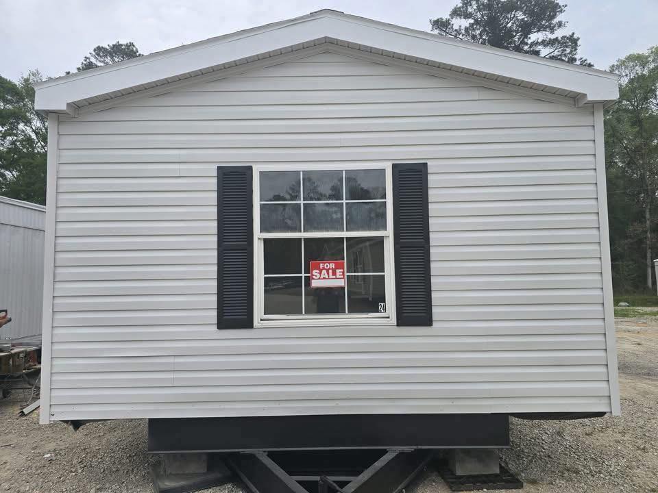 White mobile home with black shutters on display, featuring a large window and a red "For Sale" sign. The setting is outdoors with trees in the background.