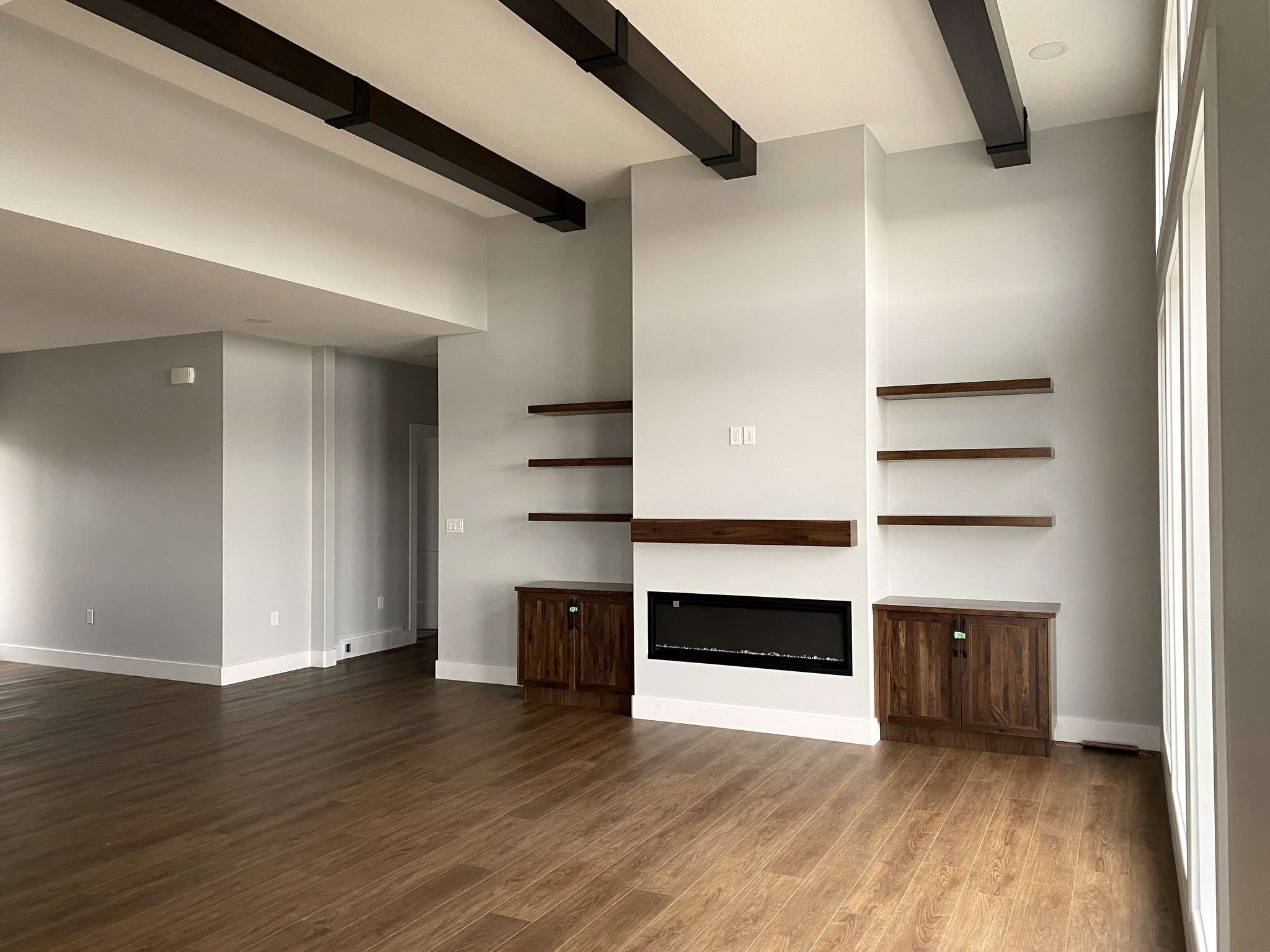 Spacious living room with light gray walls, dark wooden beams, and floor. Features include a sleek modern fireplace and floating shelves for a contemporary feel.