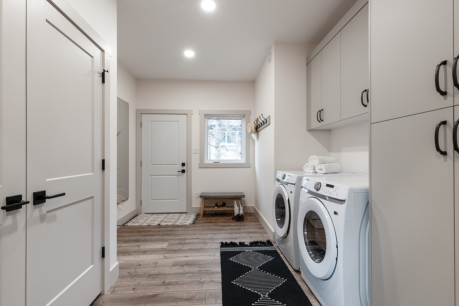 Modern laundry room with a washer and dryer, white cabinets, and wooden floor. A door and window offer natural light. The decor is minimalist.