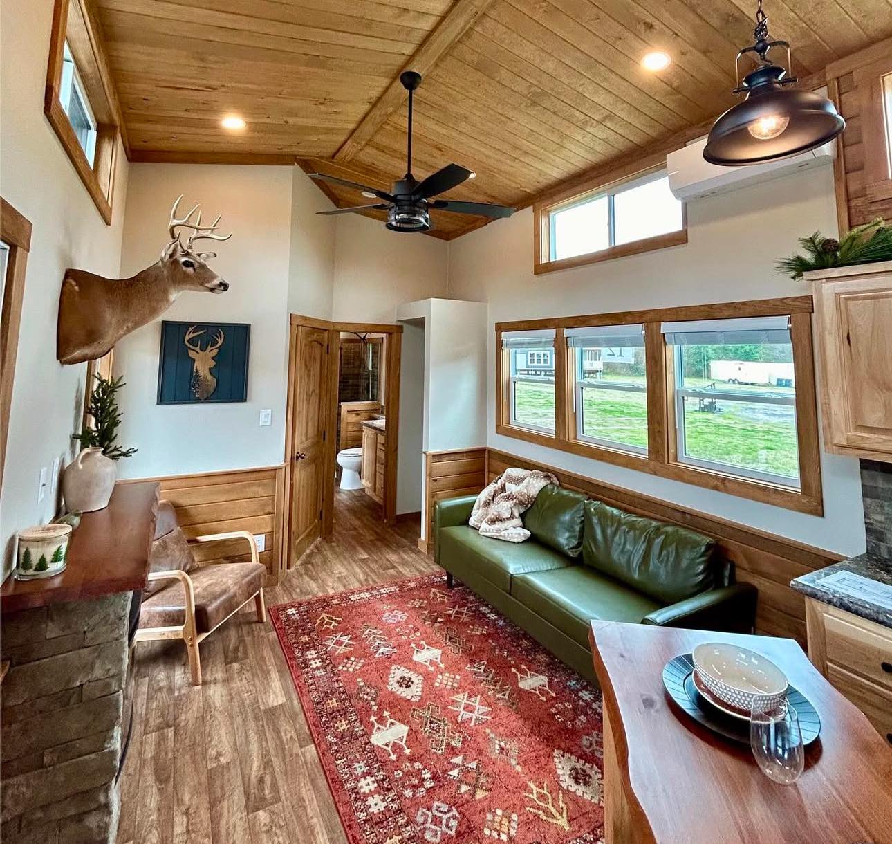 Cozy cabin interior with a vaulted wood ceiling and rustic decor. A deer head mount, green sofa, red patterned rug, and wooden accents create a warm, inviting atmosphere.