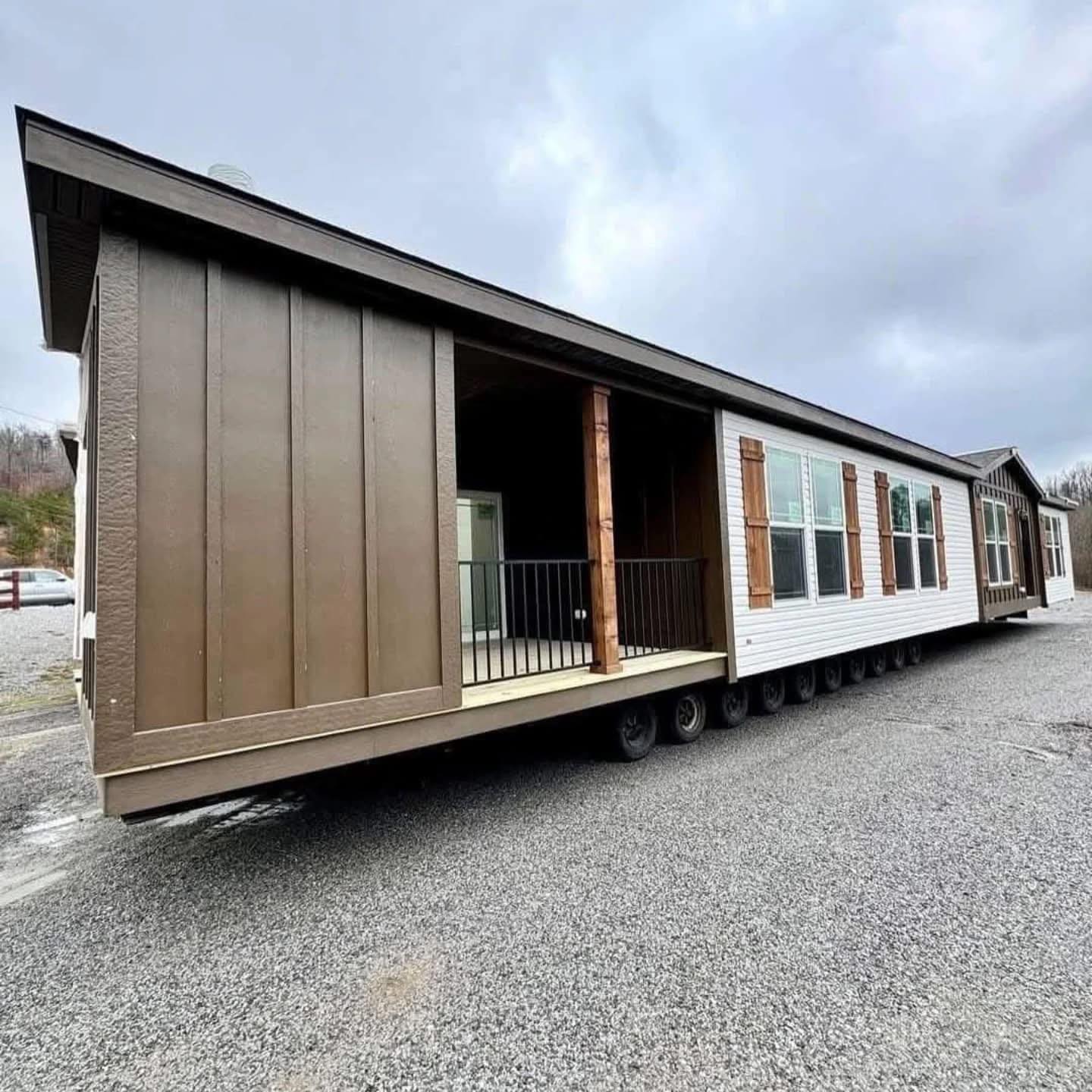 A prefab home with brown siding and white window panels stands on wheels. It has a small porch with a railing, set against a cloudy sky.