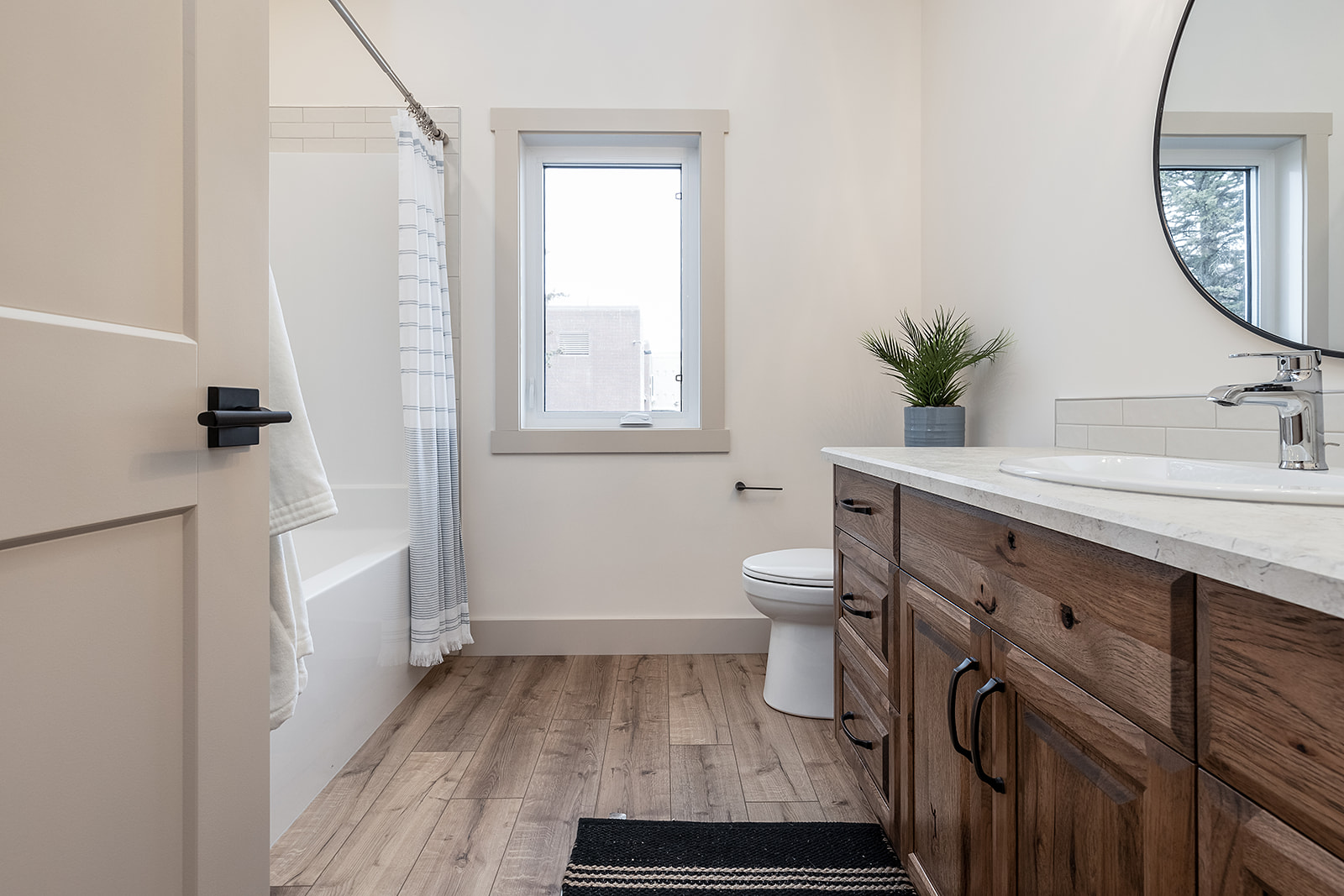 Modern bathroom with light wood flooring, a white bathtub with a striped shower curtain, wooden vanity with a white countertop, round mirror, and a potted plant.