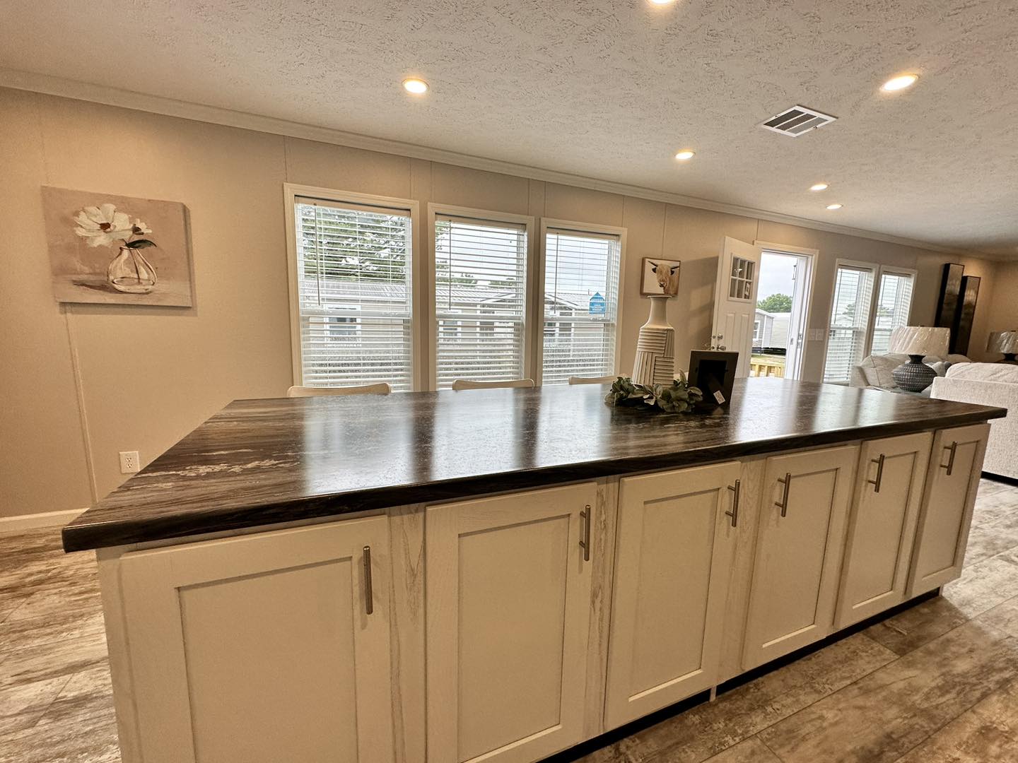 Modern kitchen interior with a large dark countertop island and white cabinets. Bright windows in the background and comfortable seating area. Cozy atmosphere.