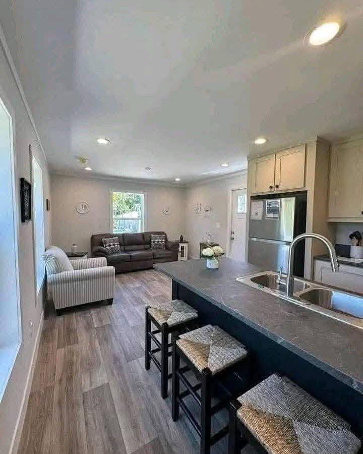 Modern open-plan kitchen and living area with wood floors. Features a dark kitchen island, stainless steel appliances, and cozy seating with neutral tones.