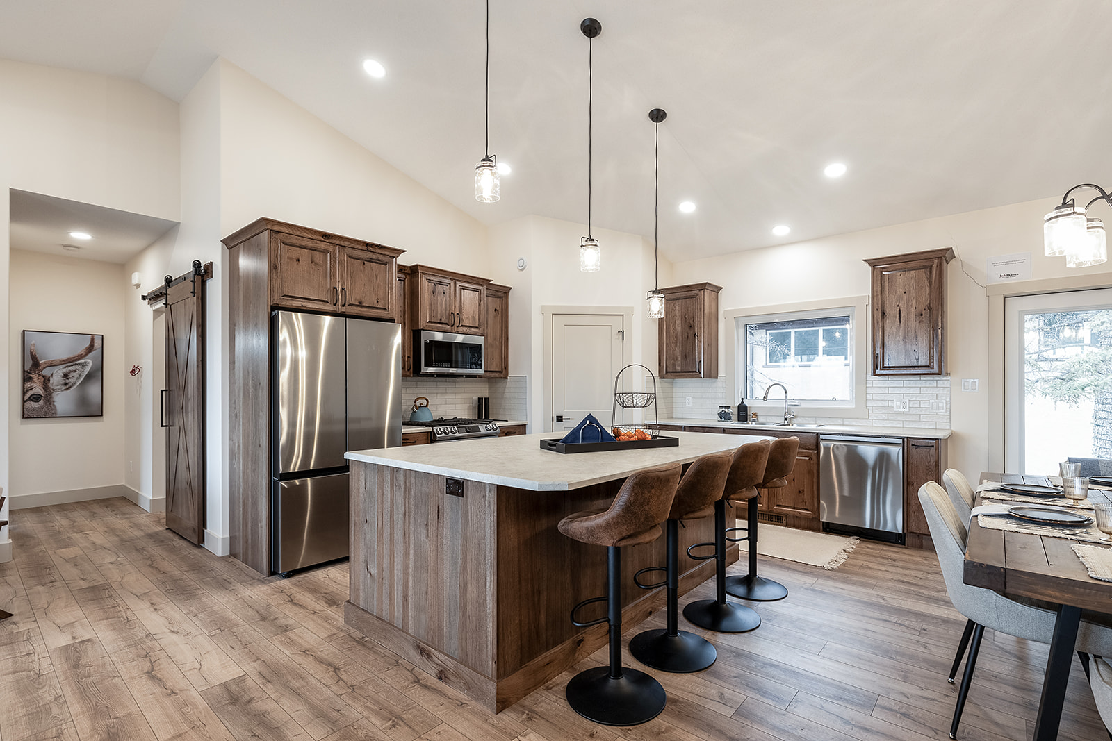 Spacious kitchen with rustic decor, featuring a large wooden island, three bar stools, stainless steel appliances, and a dining table. Warm, inviting atmosphere.