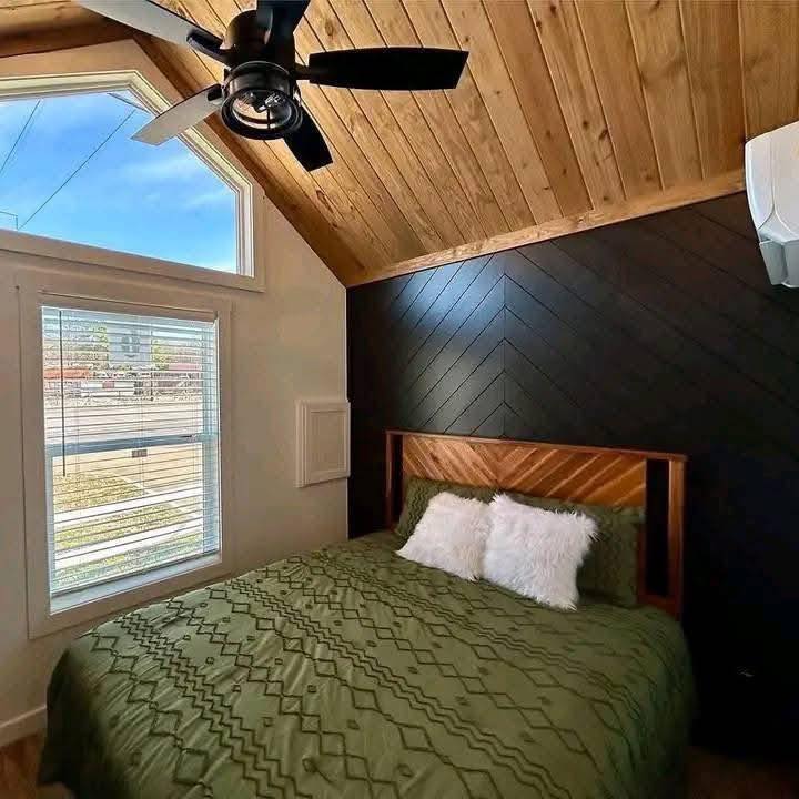 Cozy bedroom with angled wooden ceiling, ceiling fan, and large window. Green bedspread and fluffy pillows add comfort. Modern and inviting.