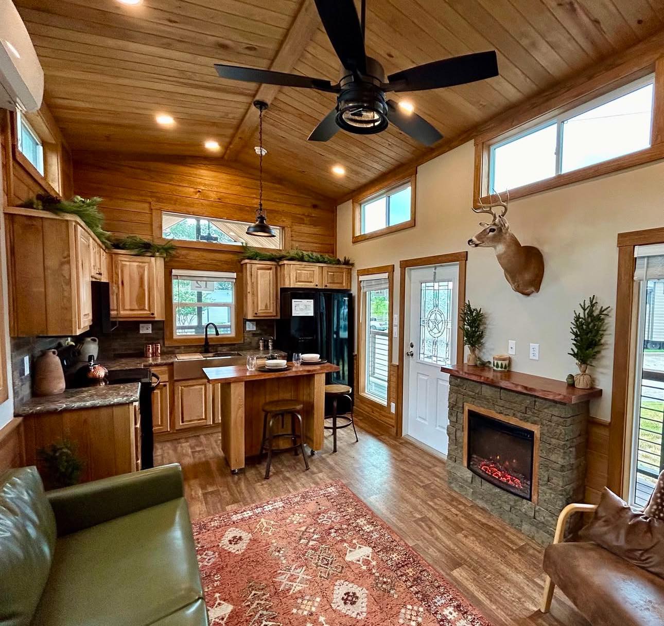 Cozy living room with a green leather sofa, rustic wood accents, and a red patterned rug. Mounted deer head and large windows create a cabin vibe.