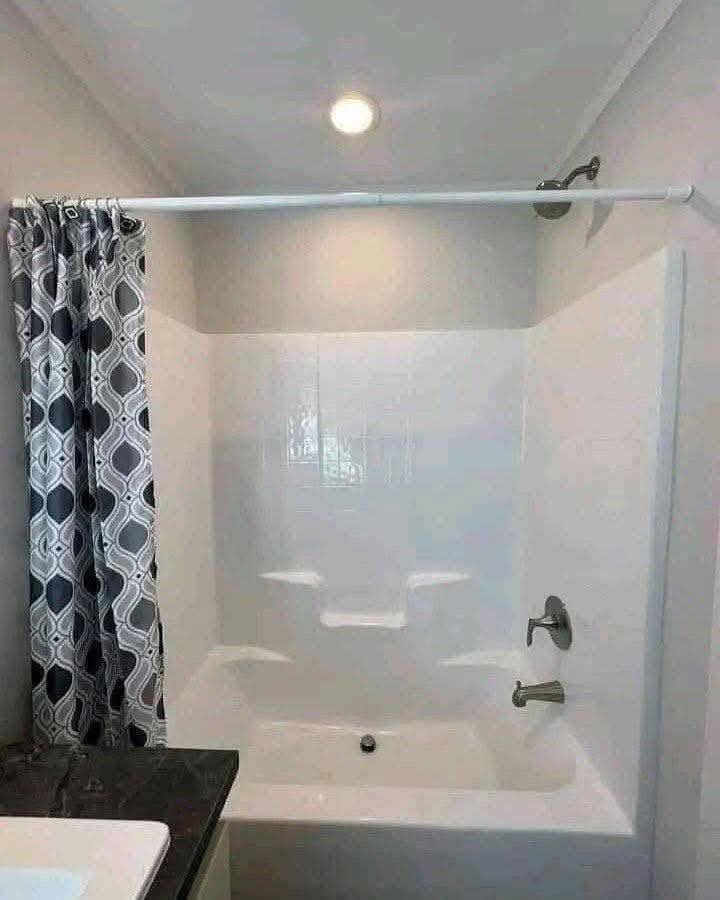 White bathtub with high walls and built-in shelves, set under a spotlight in a modern bathroom. Gray geometric shower curtain on the left adds contrast.
