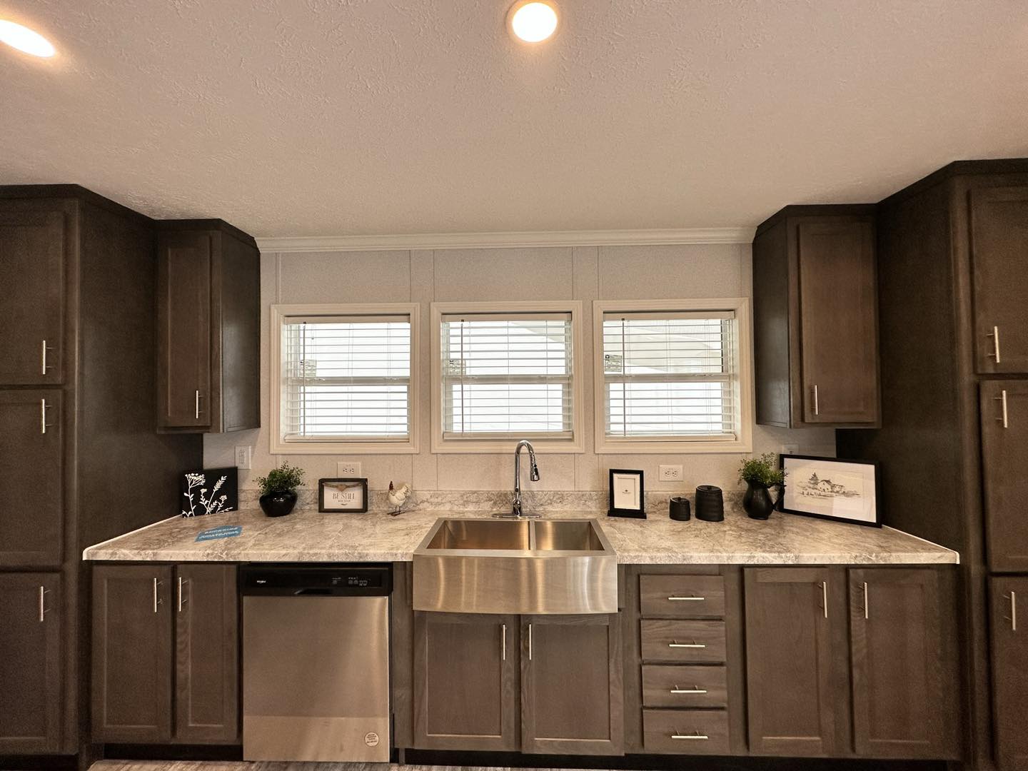 Modern kitchen interior with dark wood cabinets, marble countertops, and a stainless steel farmhouse sink. Three windows provide natural light, creating a warm ambiance.