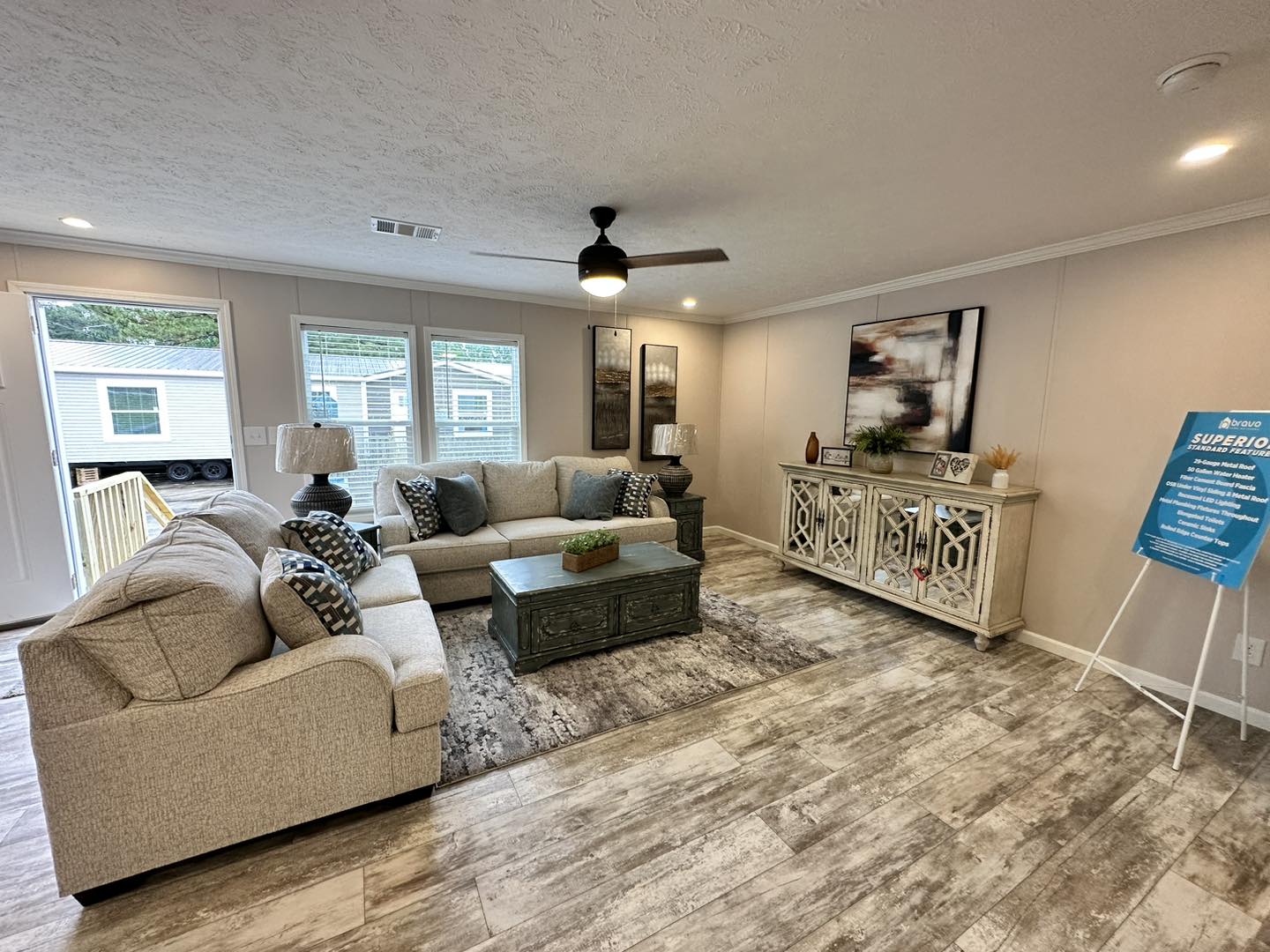 Modern living room with gray sofas, decorative pillows, and a dark coffee table on a textured rug. Artwork and lamps adorn the walls and cabinets.