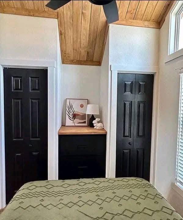 Cozy bedroom with a rustic slanted wood ceiling. Two black doors flank a dresser with art, a lamp, and decor. Olive green bedspread in view.