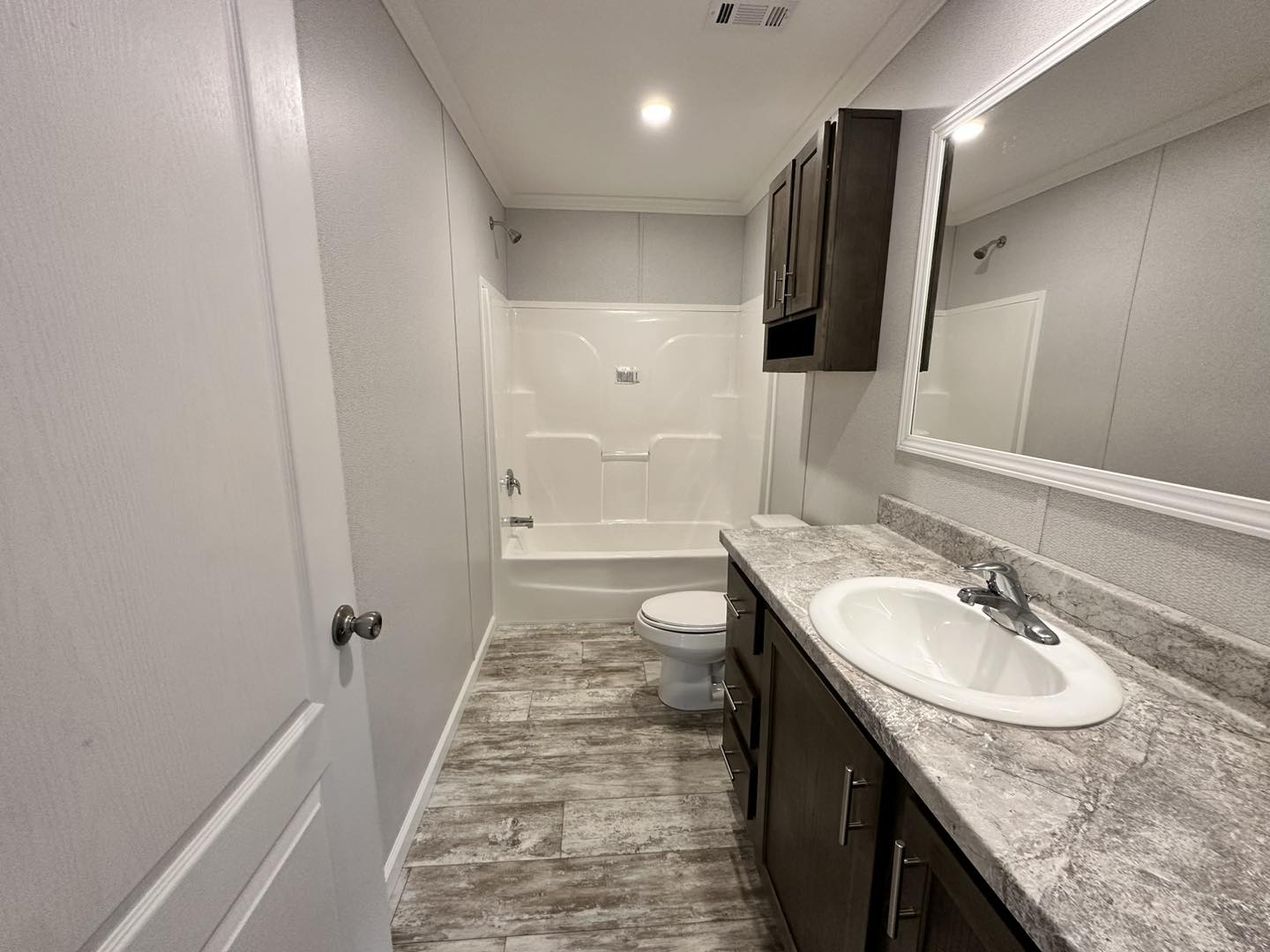 Modern, compact bathroom with gray tones featuring a sleek countertop and sink, dark vanity, toilet, and a white bathtub under bright lighting.