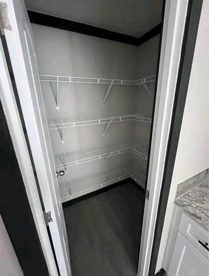 Empty pantry closet with three wire shelves on two walls, gray flooring, and open white door. Adjacent to kitchen countertop and cabinet.